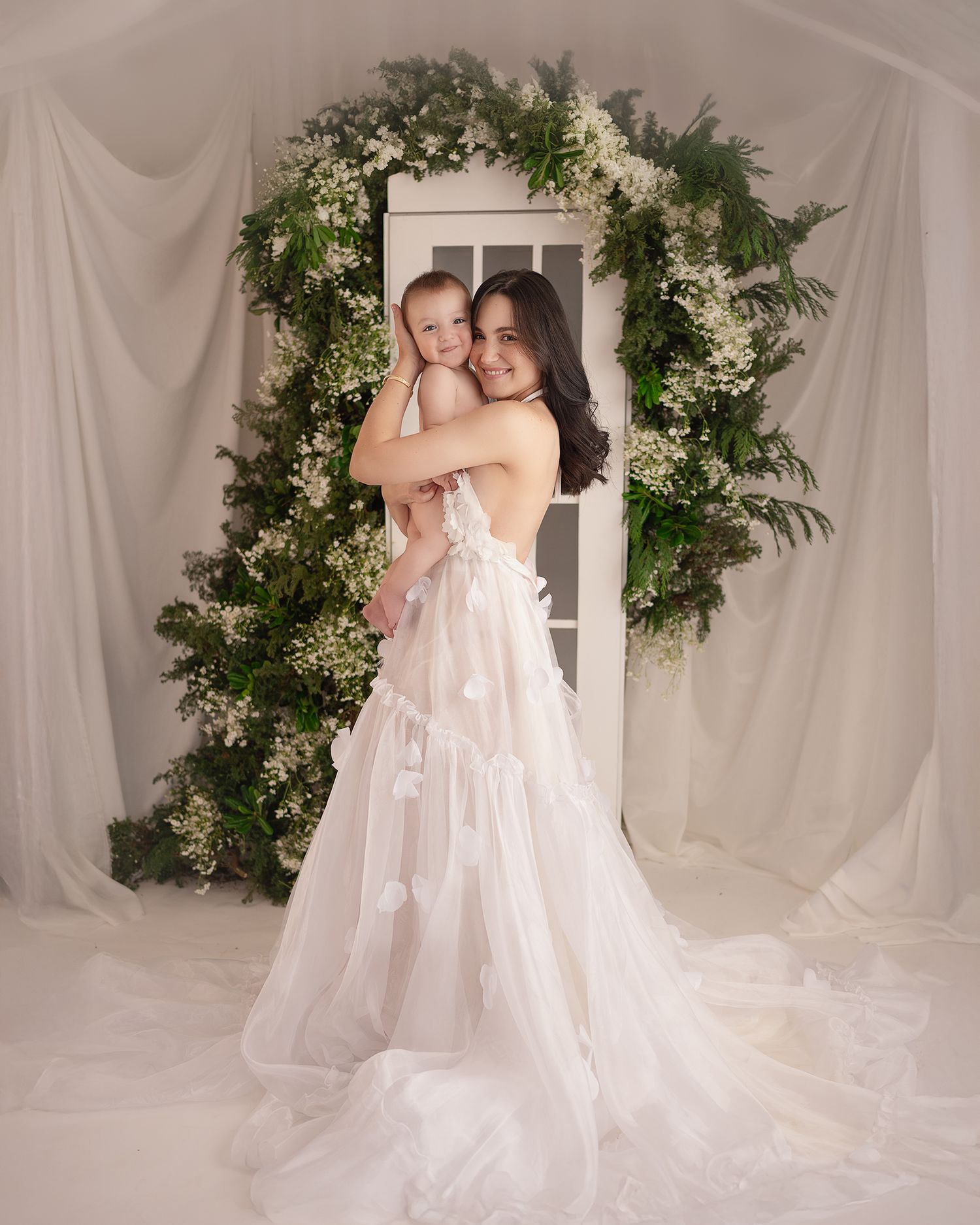 Elegant white studio setup with draped fabric and greenery arch for portraits.