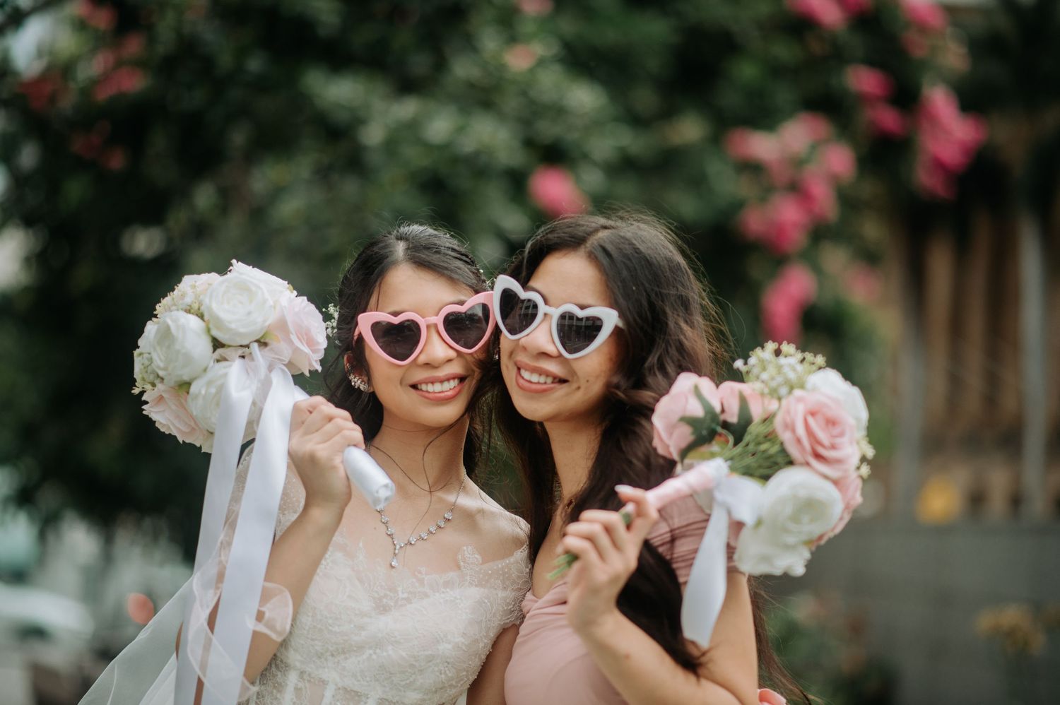 Two wedding guests in white dresses posing with bouquets and heart-shaped sunglasses against a floral backdrop.