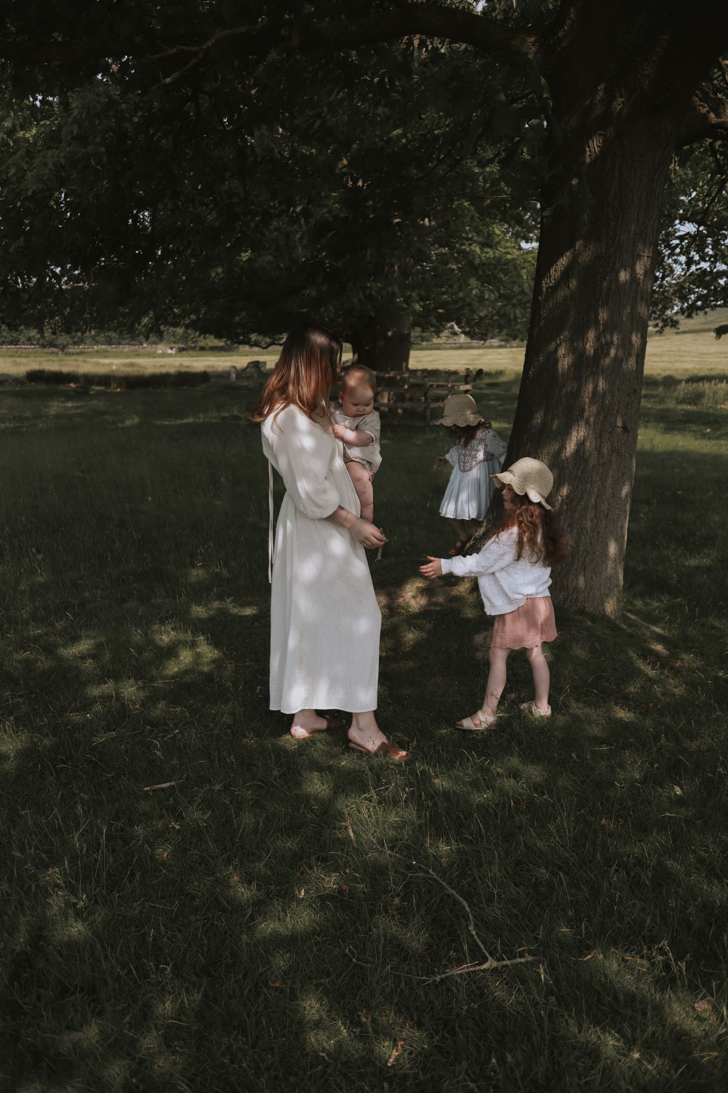 A mother and her children in white dresses share a tender moment under a shady tree in a park setting.