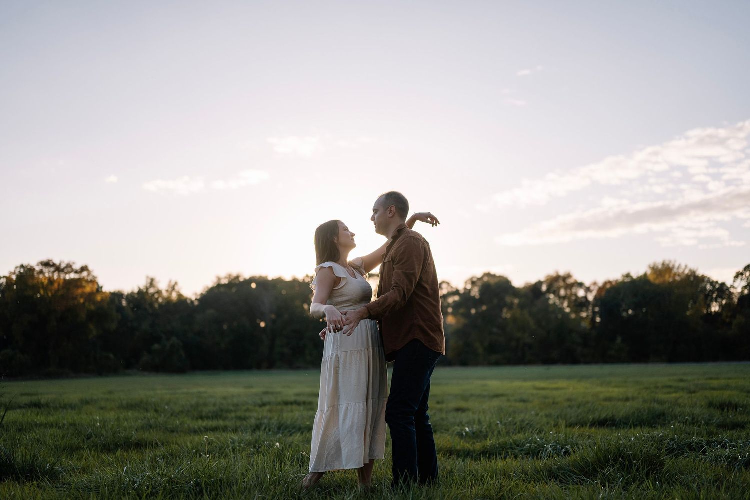 Romantic couple standing close in meadow during golden hour with white dress and casual wear.