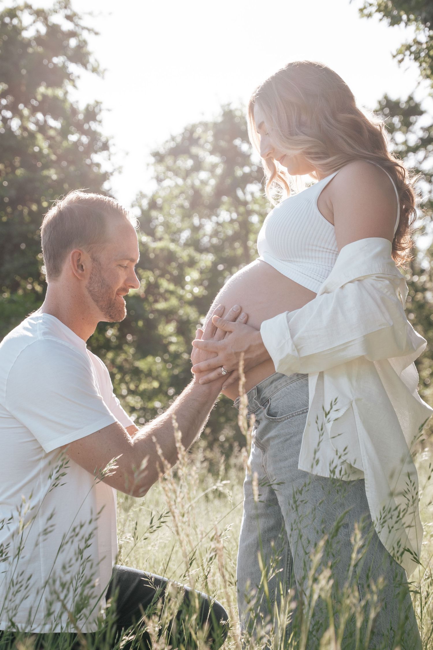 Man kneels holding pregnant partners belly in sunny outdoor field with tall grass.