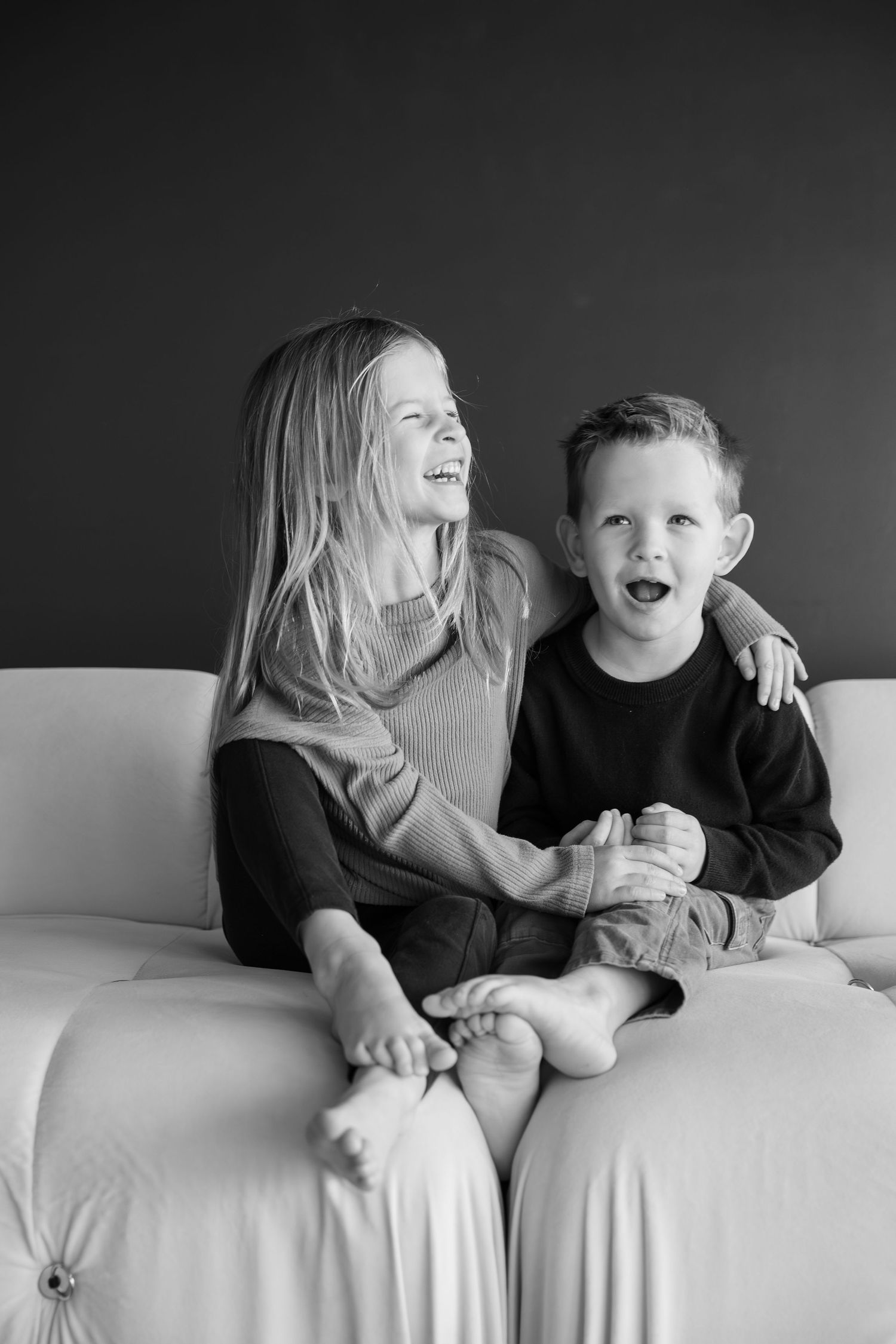 Black and white photo of two happy children, a girl and boy, laughing together on a white couch in studio photoshoot