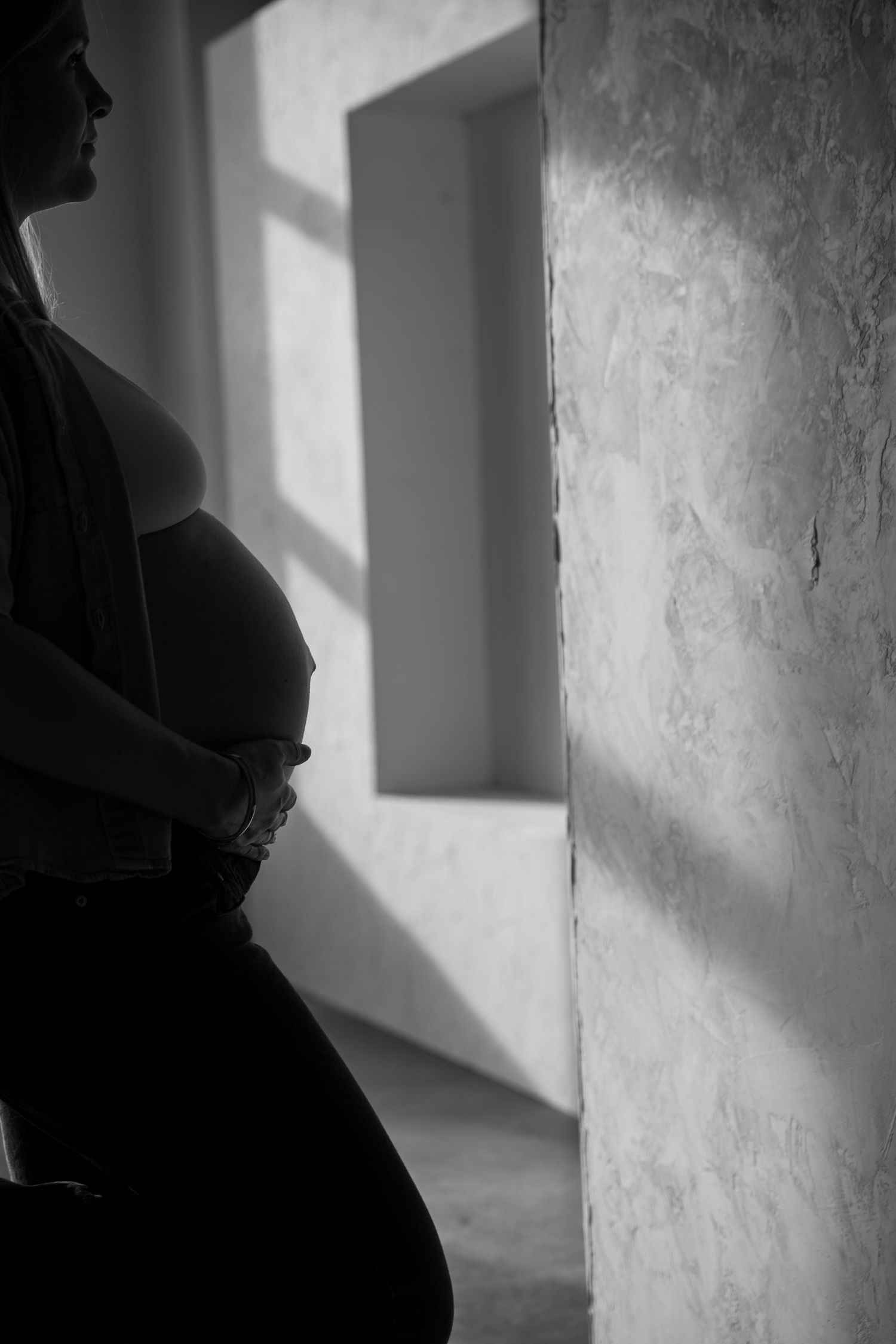 Black and white silhouette of a pregnant woman cradling her baby bump near a sunlit window.