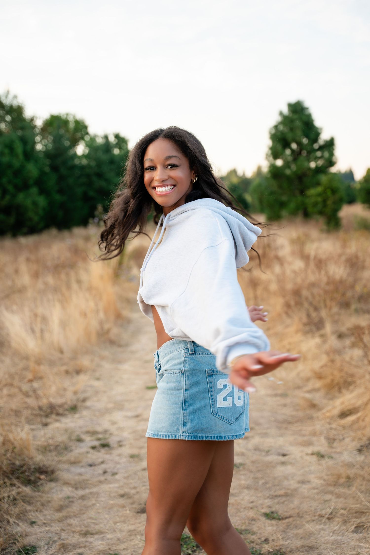 Smiling young woman in white hoodie and denim mini skirt posing on a rural path surrounded by golden grass.