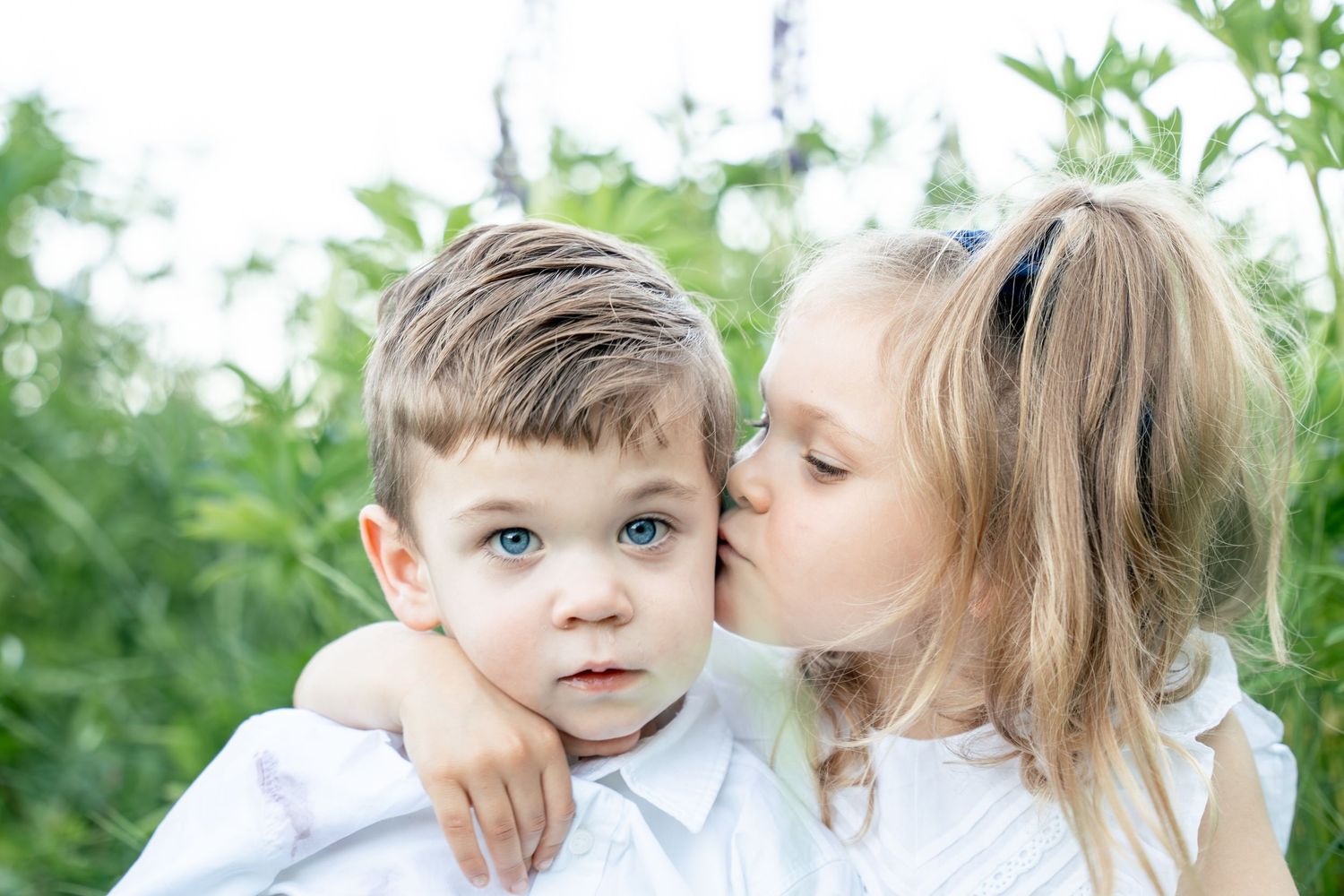 Young girl kisses boy on cheek outdoors surrounded by green foliage.