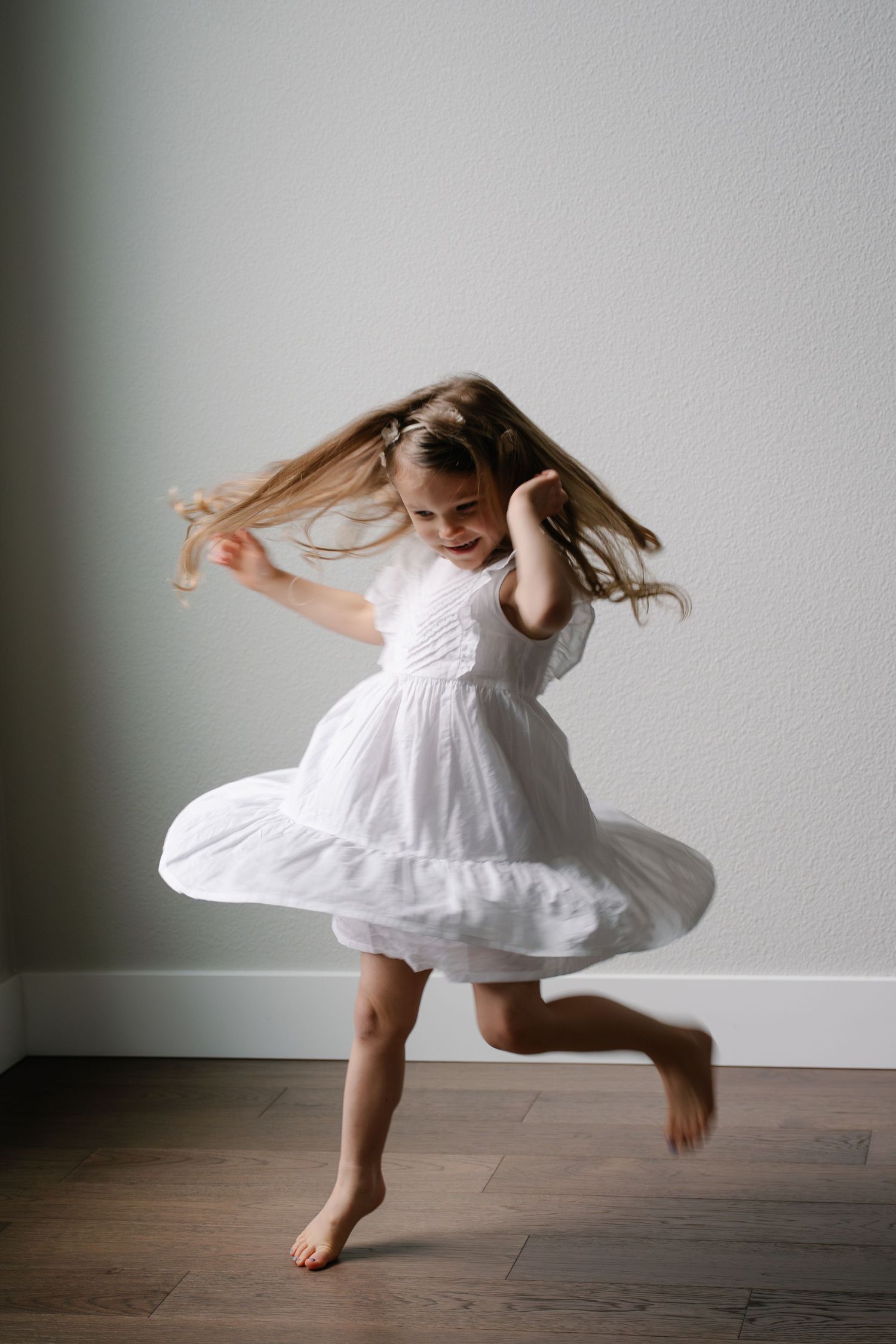Joyful little girl in white dress twirling and dancing barefoot on hardwood floor.