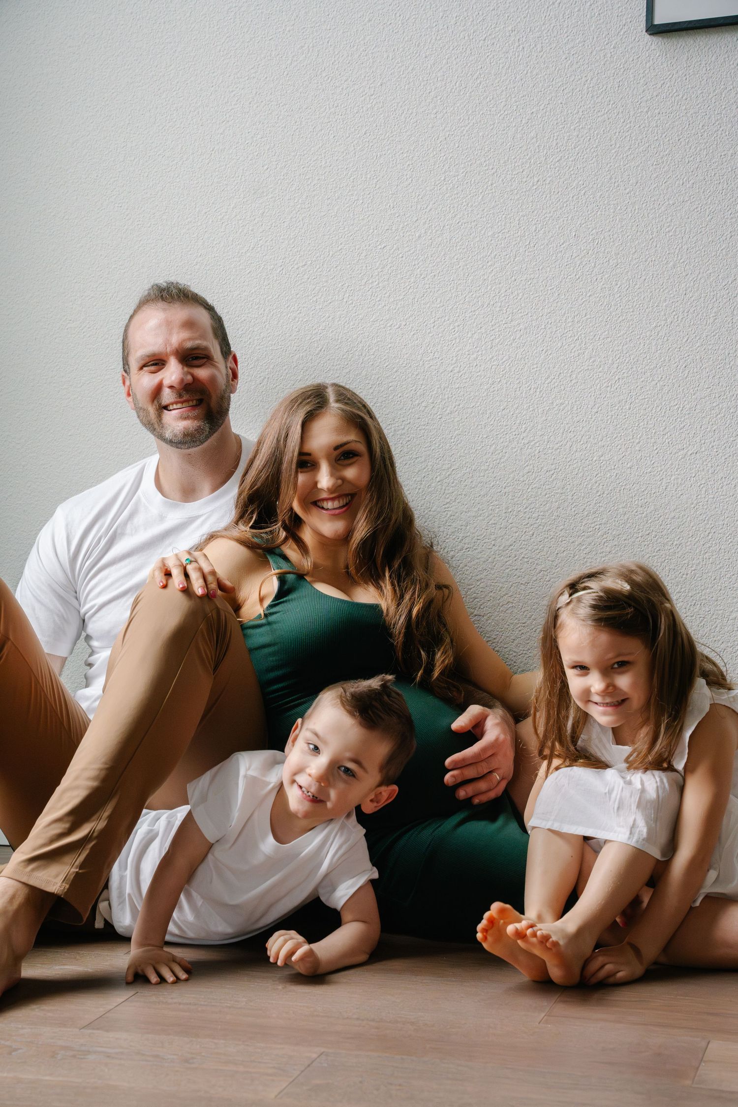 Happy family of four sits casually on the floor together against a white wall.