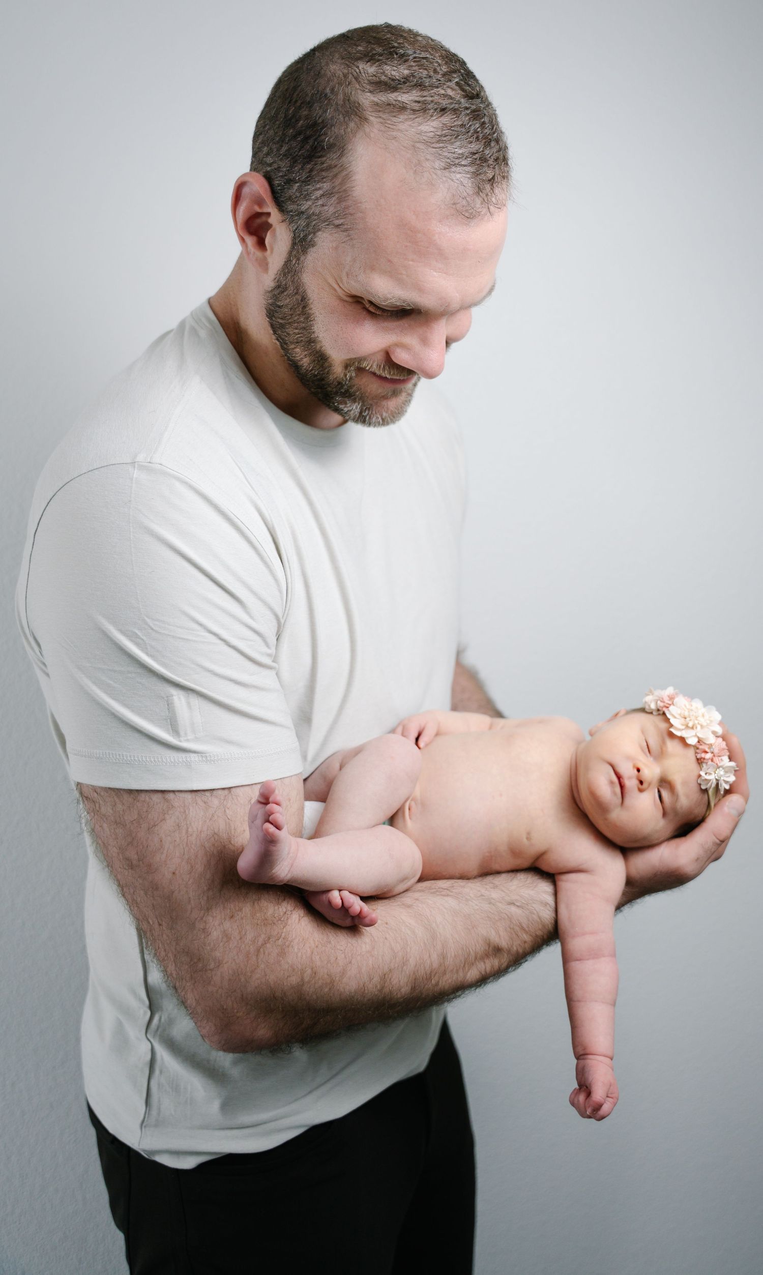 Bearded father in white t-shirt smiles at sleeping newborn baby with floral headband.