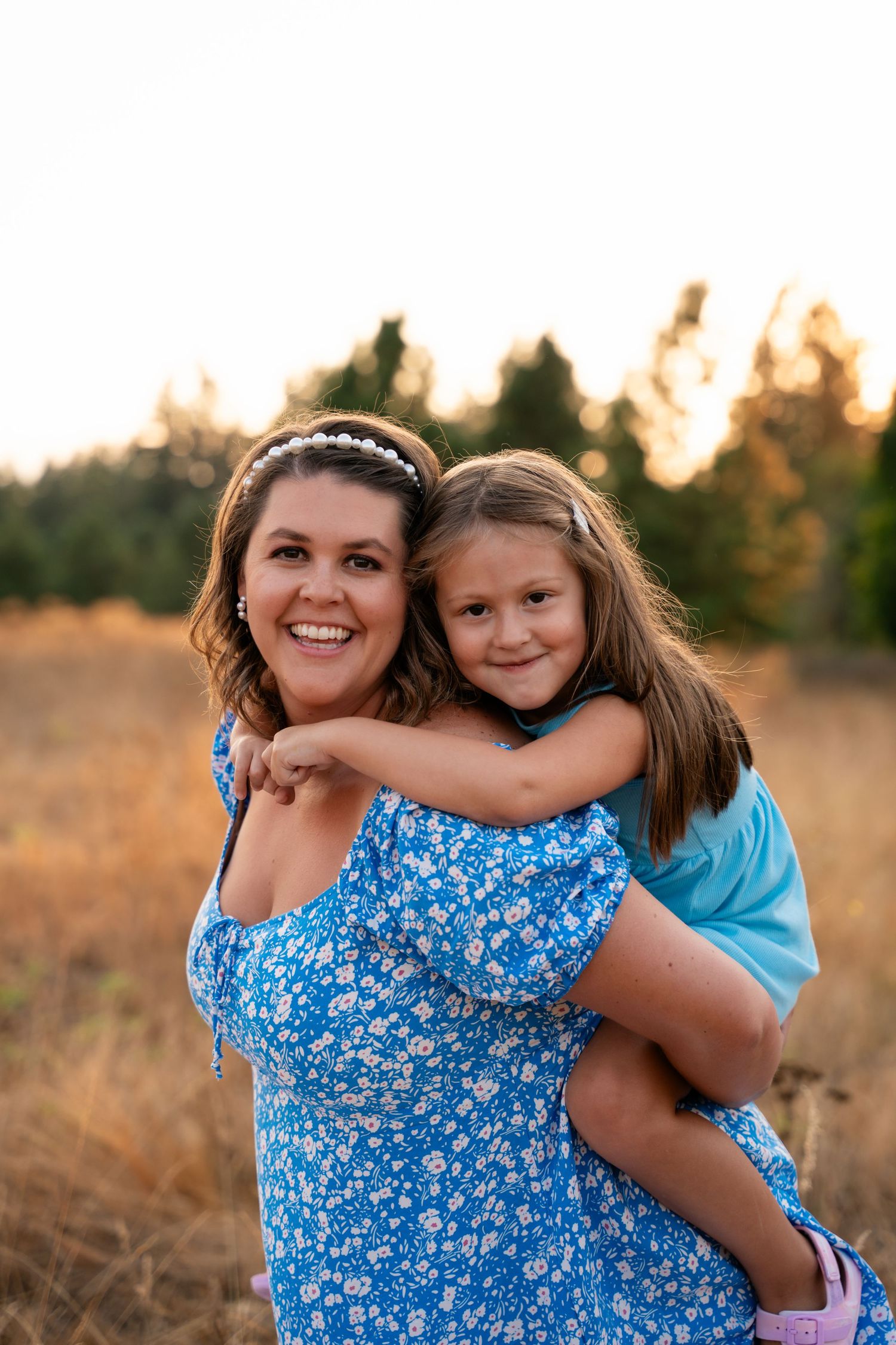 A smiling woman in a blue floral dress gives a young girl a piggyback ride in a sunny golden hour photoshoot