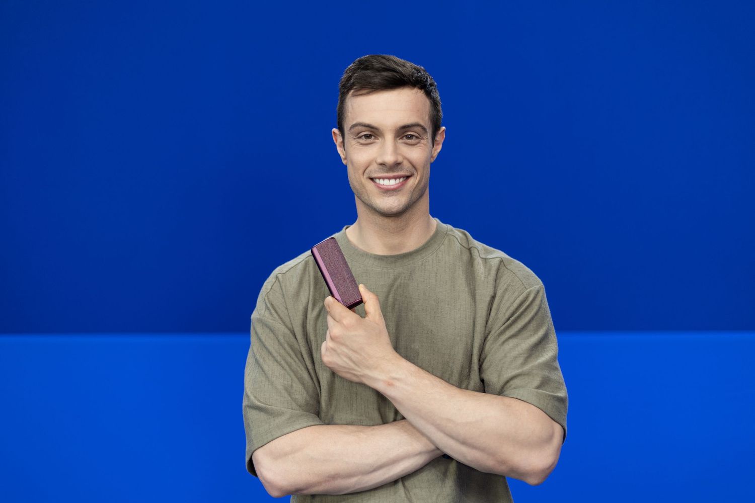 Smiling young man in olive t-shirt holding a book with arms crossed against a bright blue background.