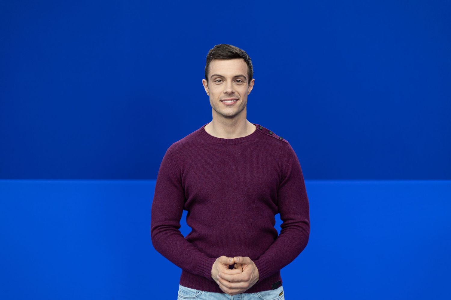 Young man in burgundy sweater smiling against a blue background, standing with hands clasped.