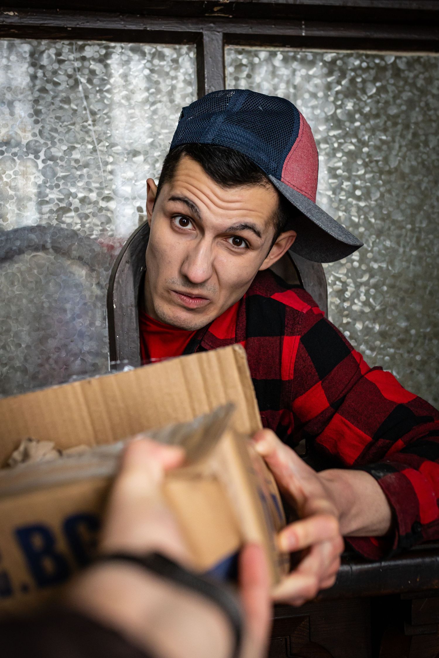 Young man in red plaid shirt and snapback cap receiving a cardboard box through a window.