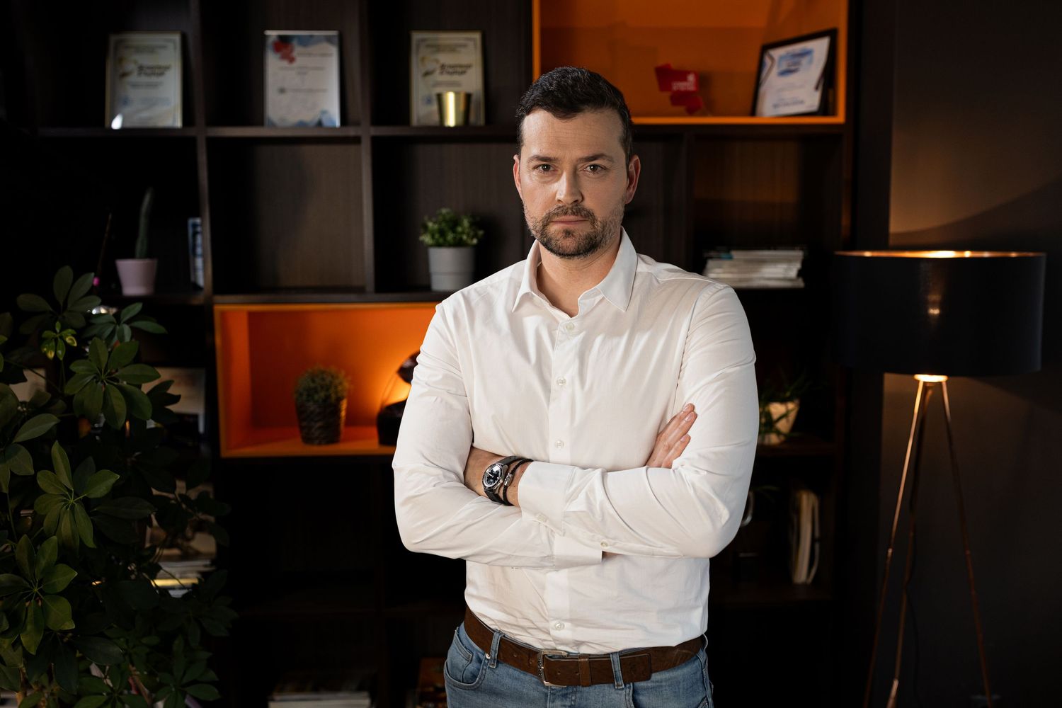 Professional man in white shirt stands confidently with arms crossed in a modern office setting.