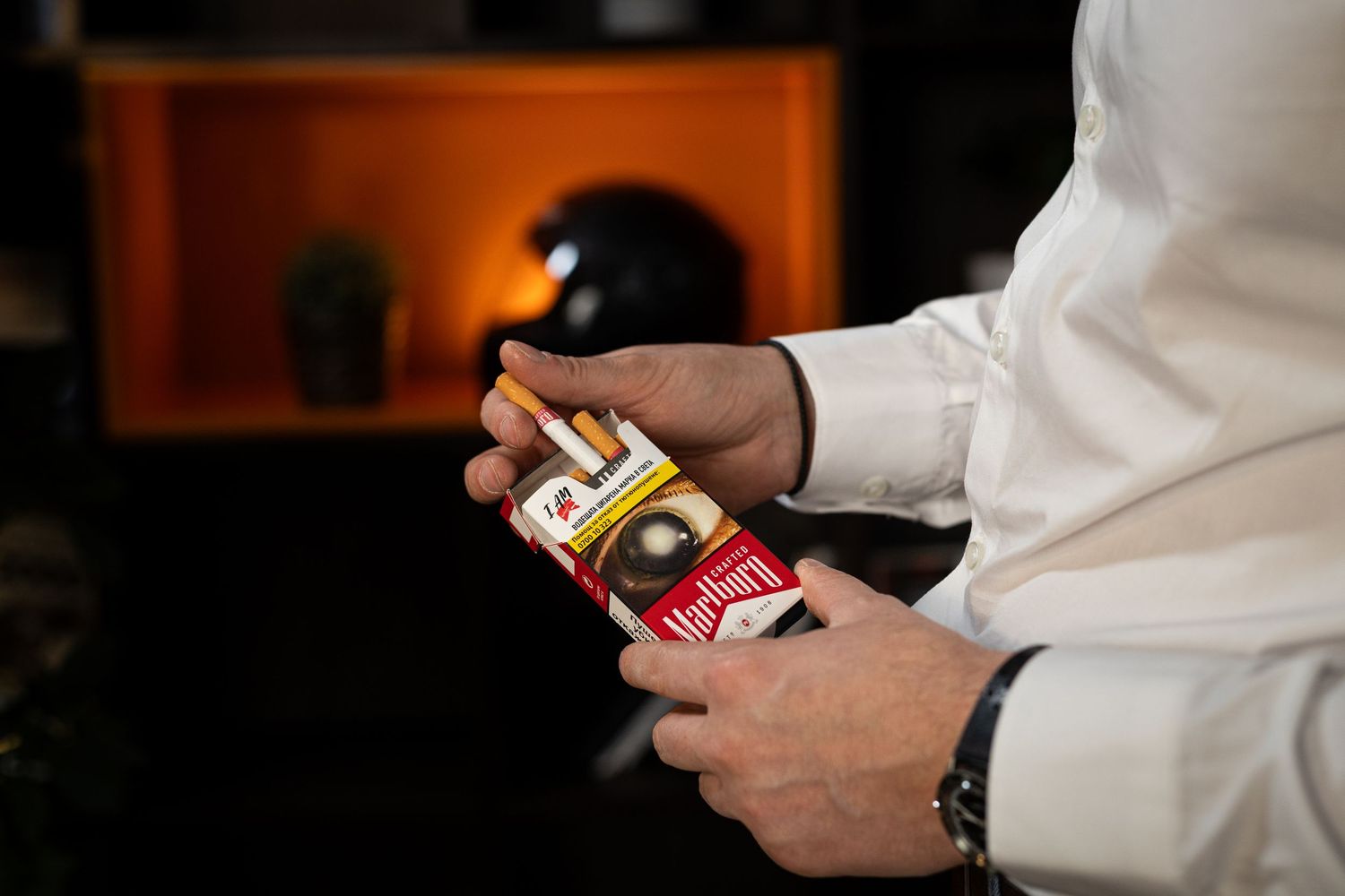 Man in white shirt opening a red Marlboro cigarette pack, with a dark blurred background.