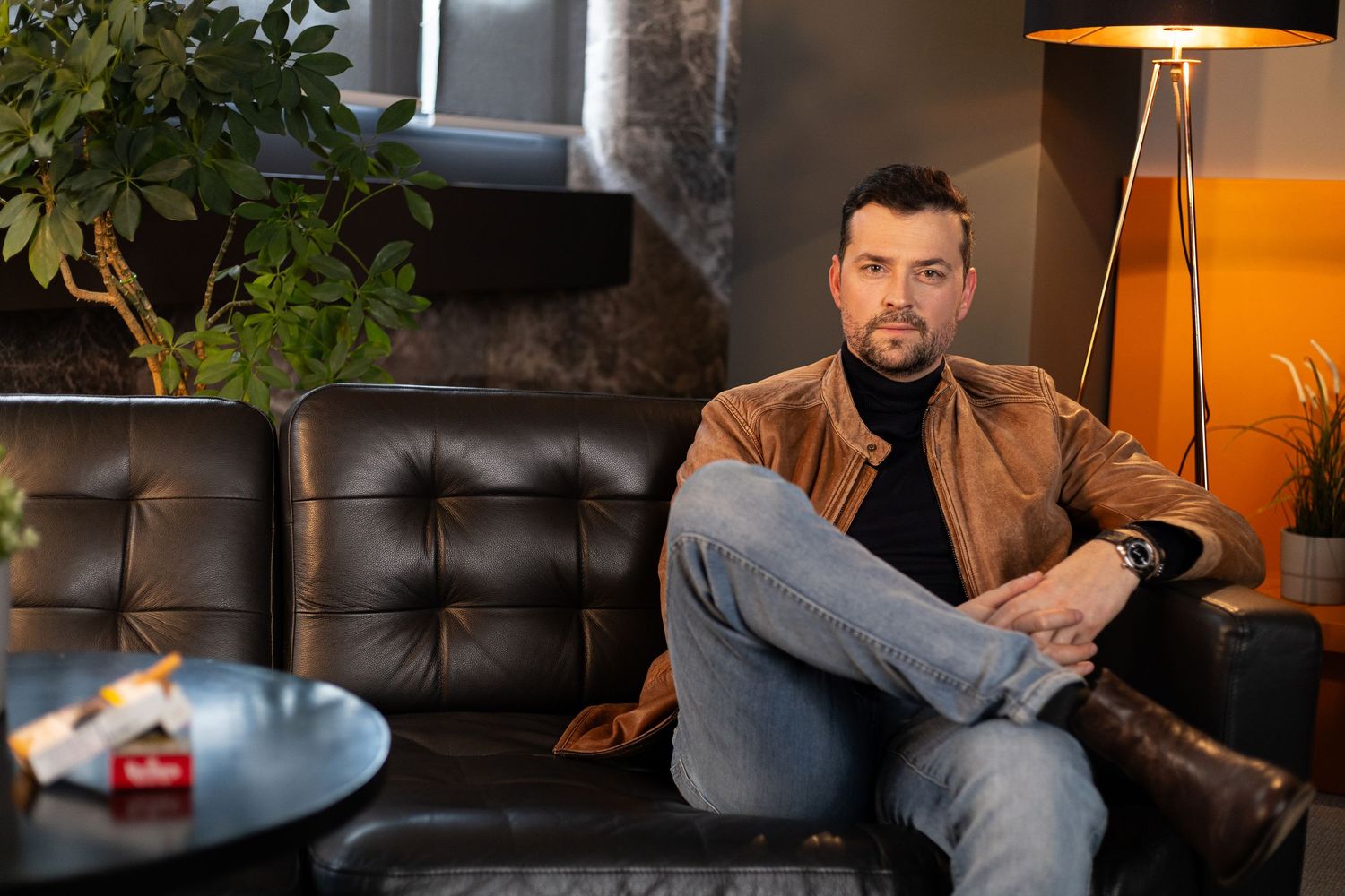 Man in brown leather jacket and jeans sitting relaxed on dark leather sofa in modern interior.