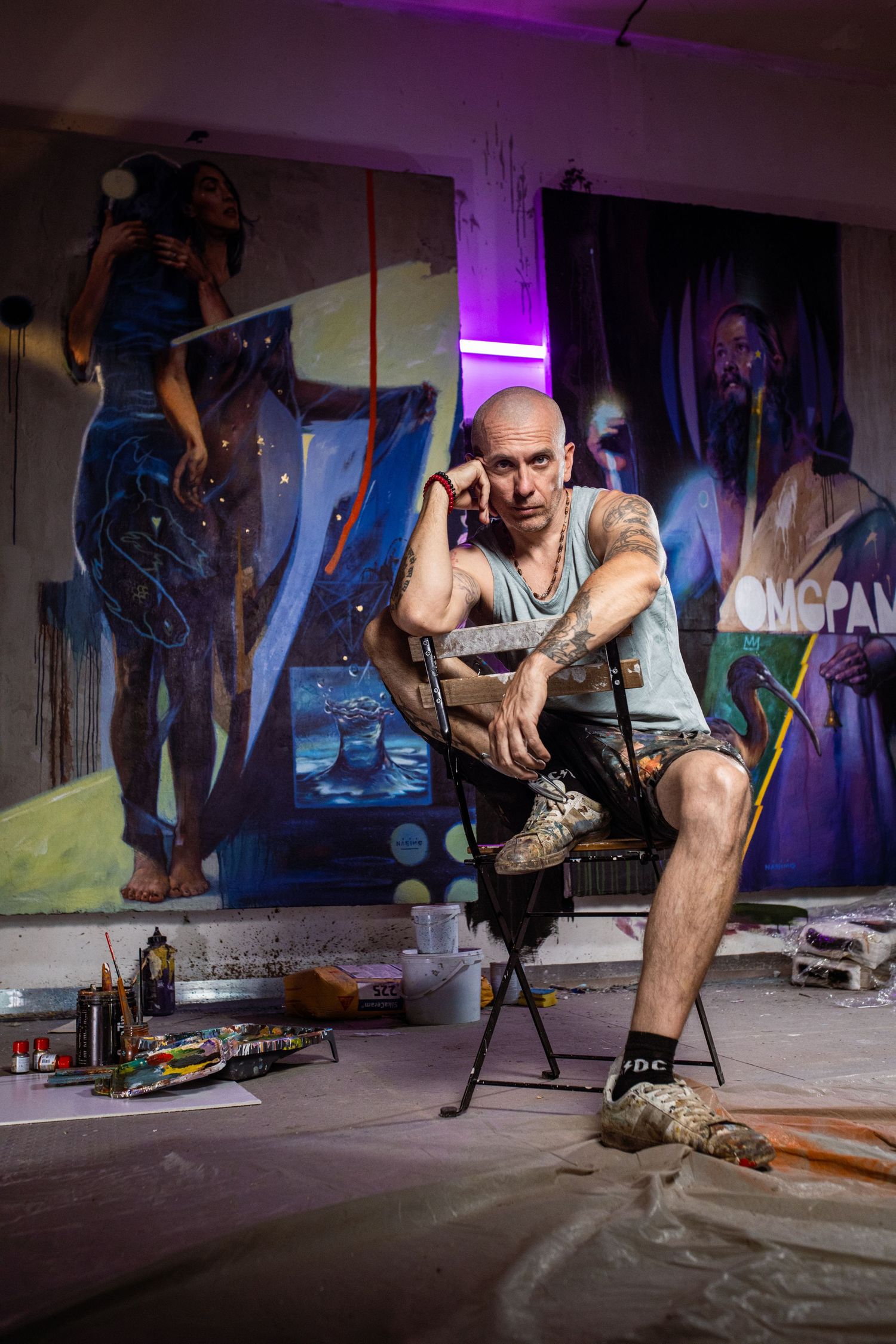 Bald tattooed artist sitting in studio surrounded by large figurative paintings with art supplies on floor.