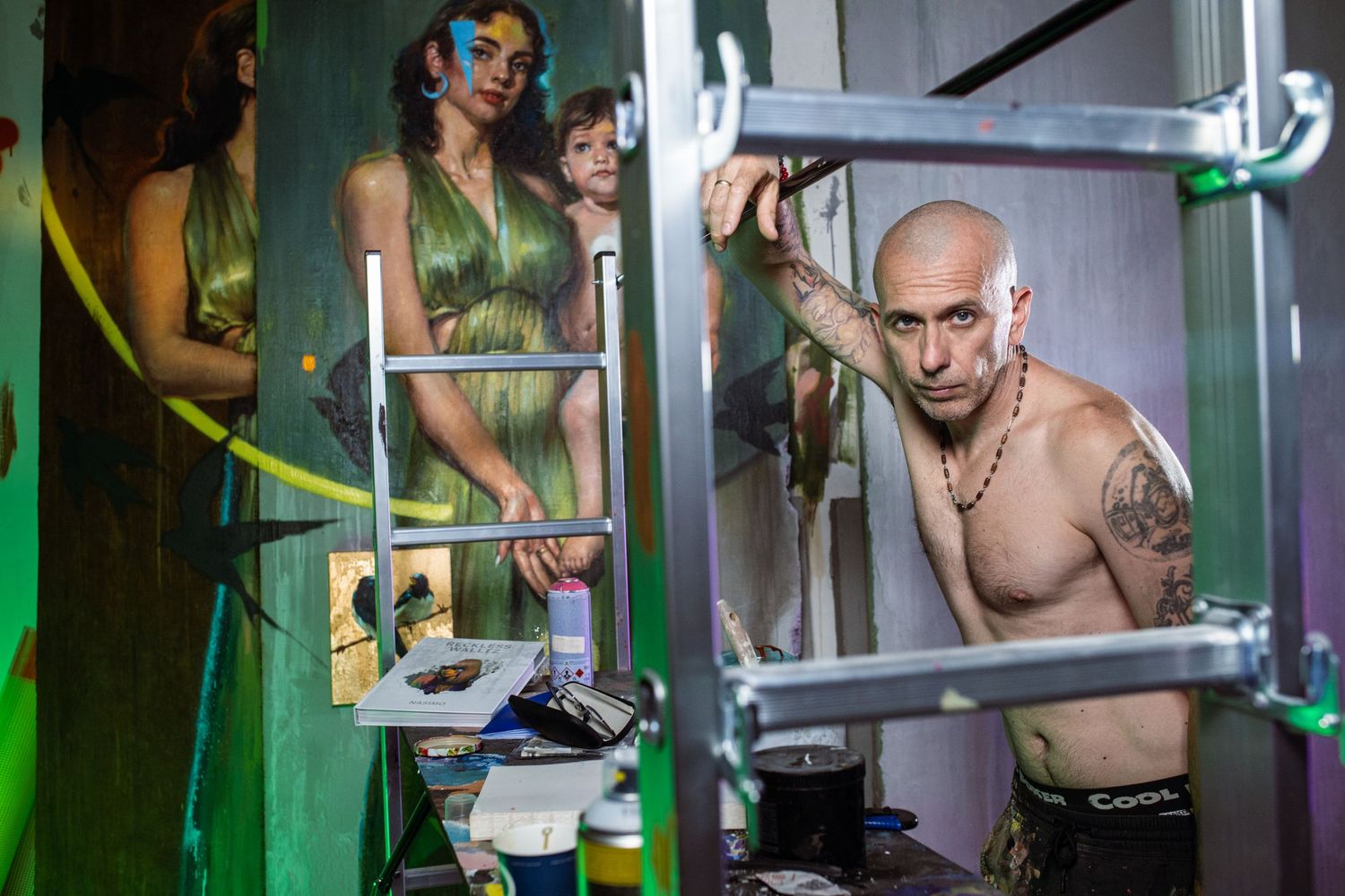 Bald tattooed male artist works on body painting project with painted model in background.