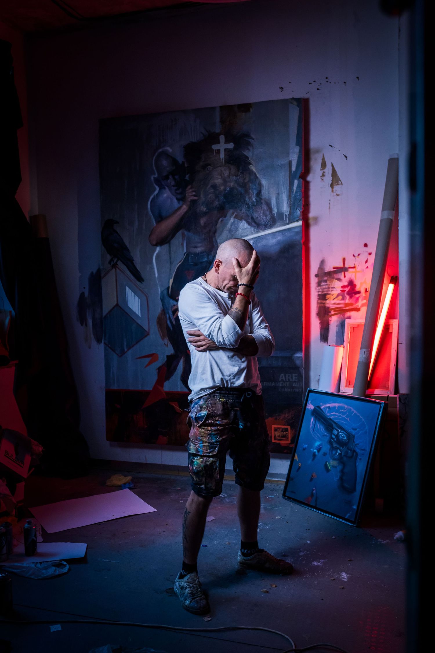 Tattooed bald artist covering face with hands standing in dark studio with large paintings and red neon light.