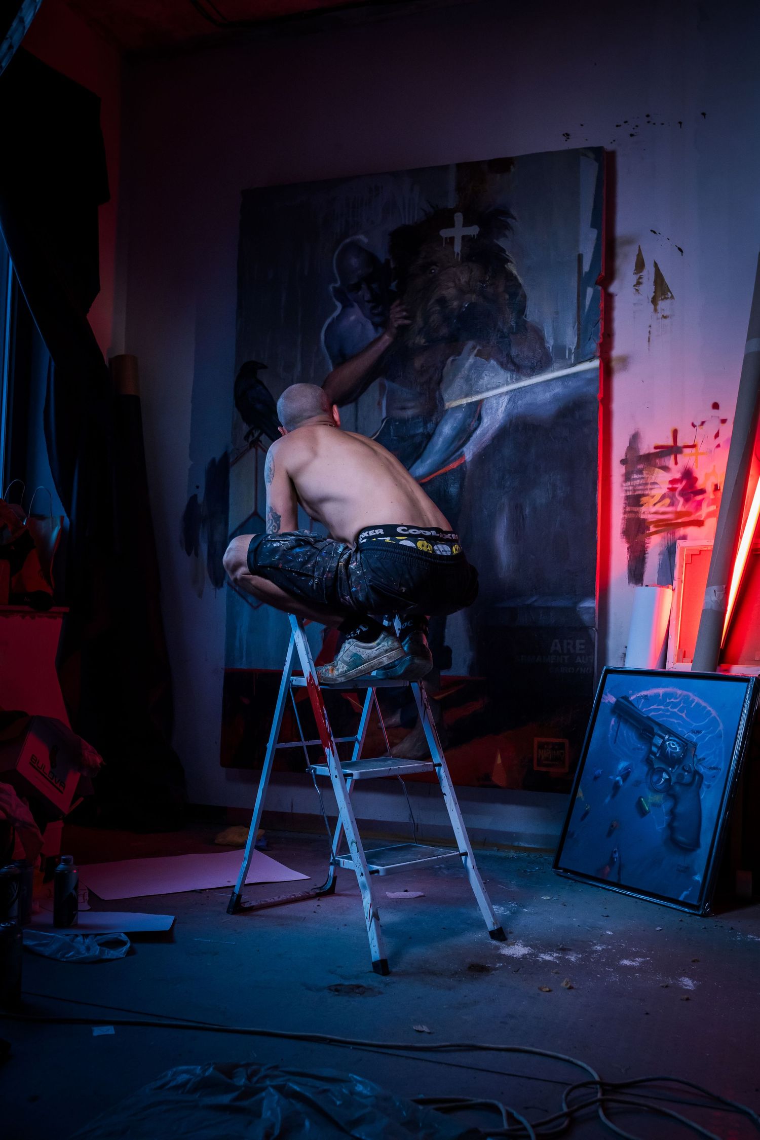Shirtless tattooed artist on ladder painting large dark canvas in moody blue and red lit studio.