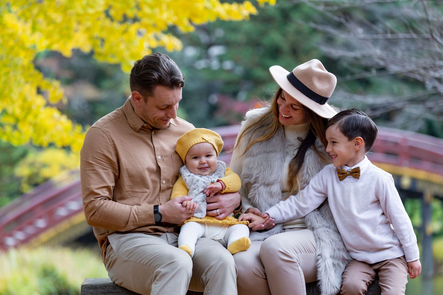 A family in coordinated neutral outfits sits together outdoors on a bench during autumn with yellow foliage in background.