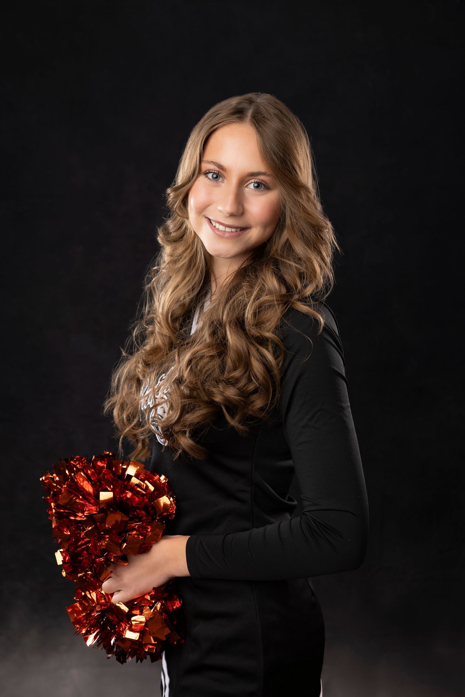 Cheerleader in black outfit holds red and gold pom poms against dark background.