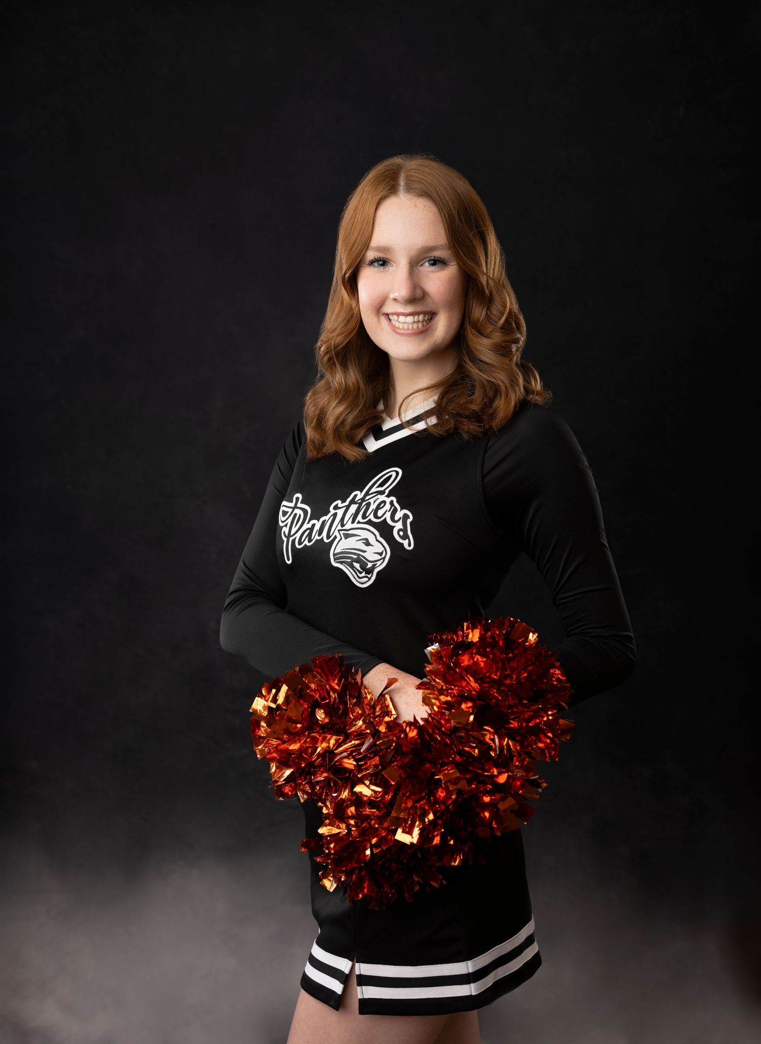 Cheerleader smiles while holding red pom poms in black and white uniform.