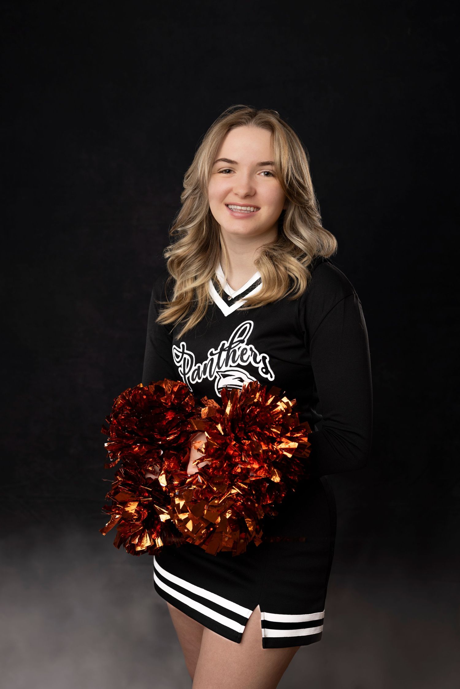Cheerleader in black uniform poses with red pom poms against dark backdrop.