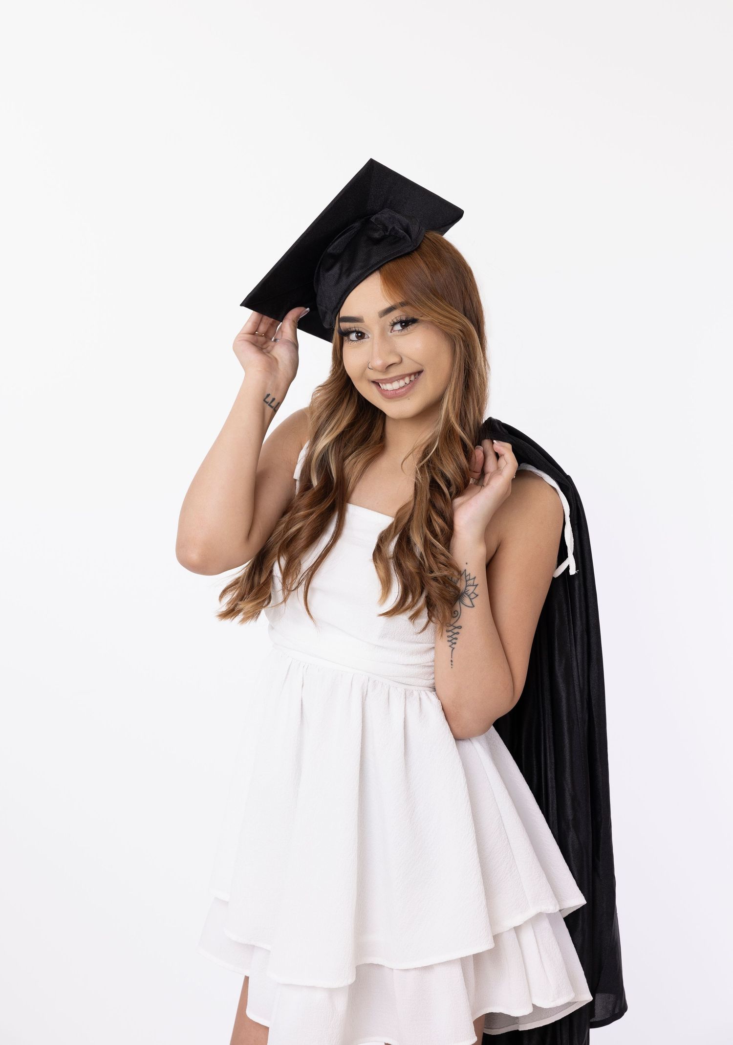 Graduate in white dress holds graduation cap against white background.