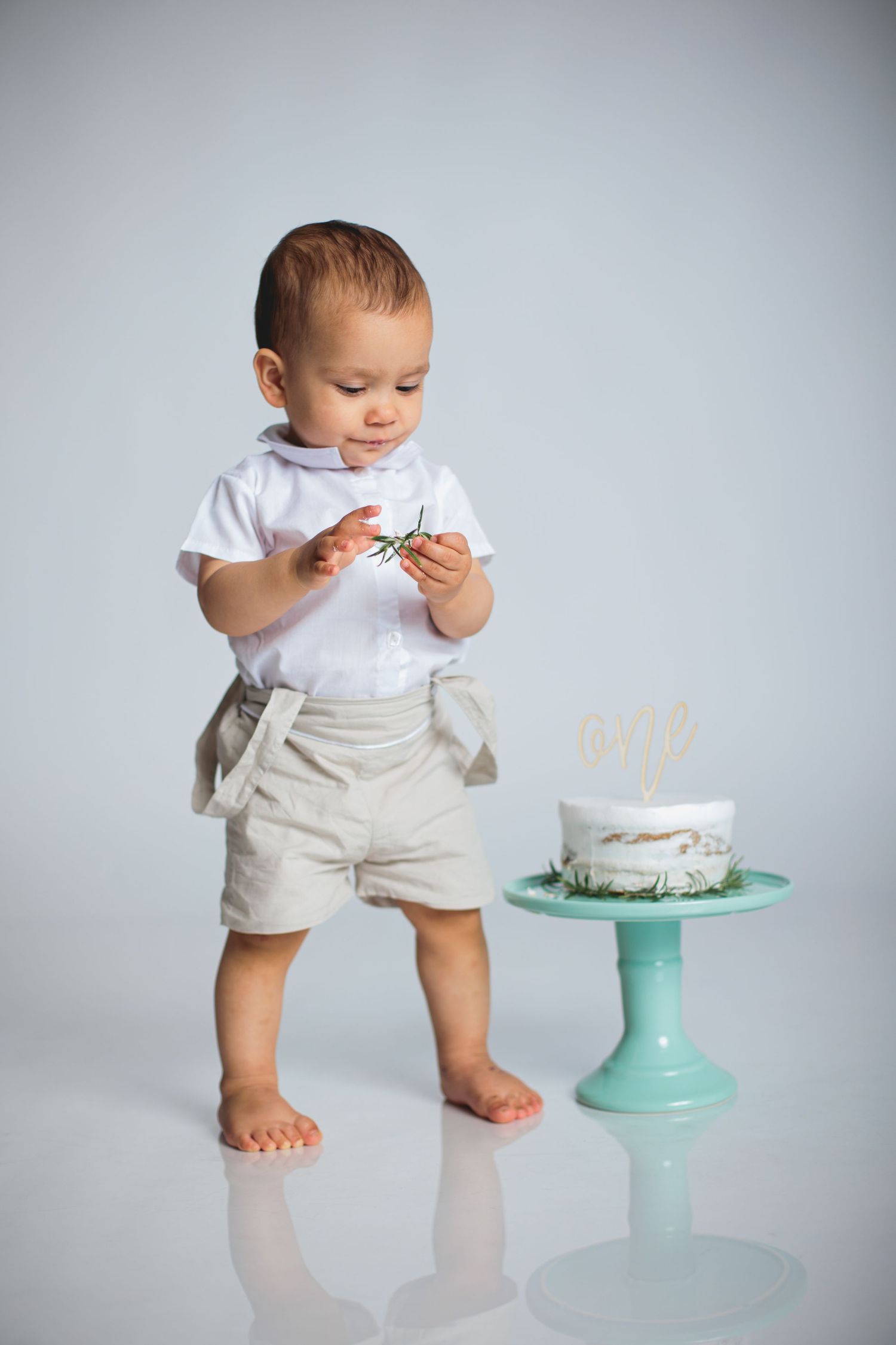 Small child in white shirt and khaki shorts stands barefoot next to birthday cake on mint green cake stand.
