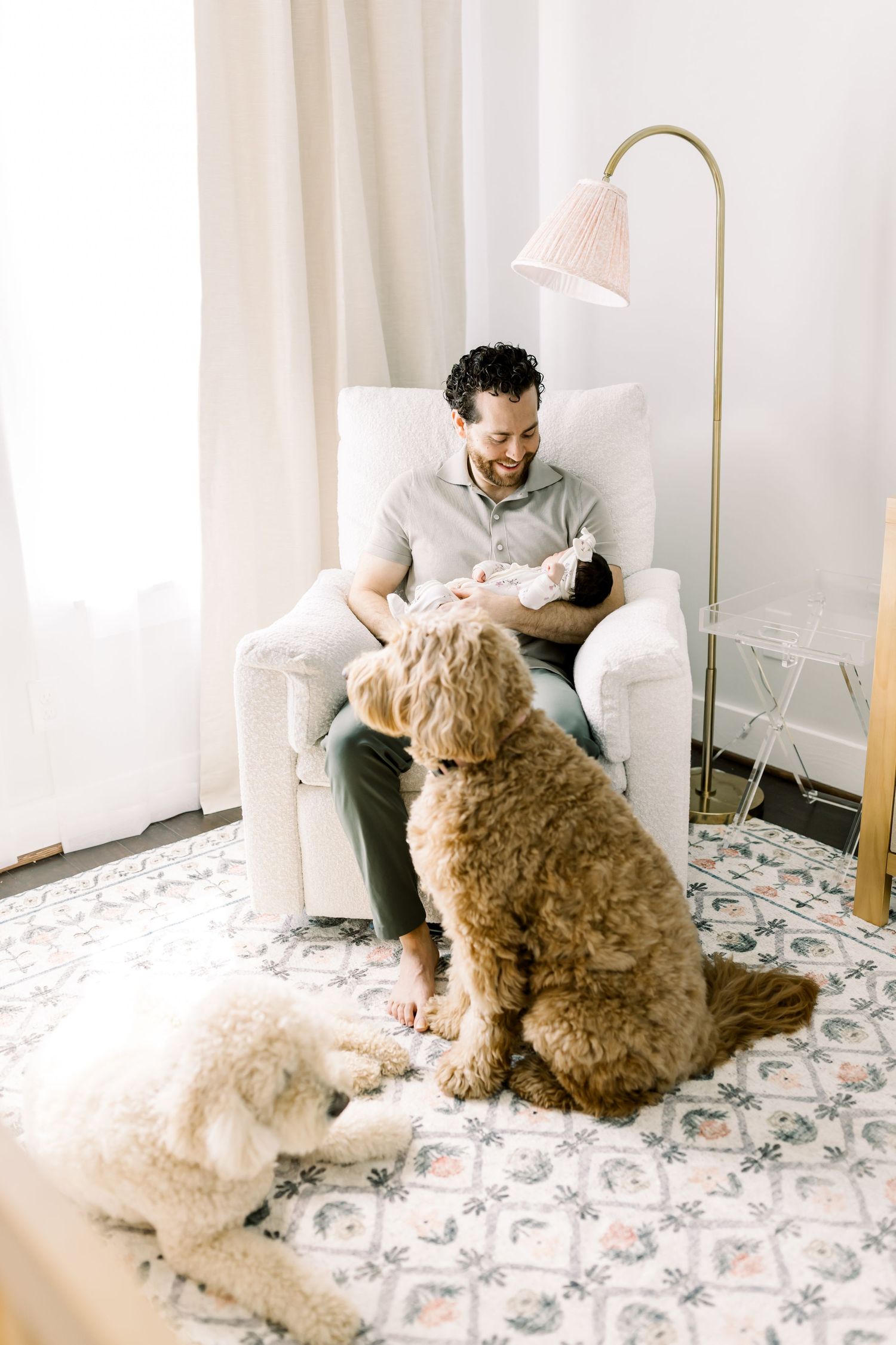 Two goldendoodles sitting attentively near a white armchair under a floor lamp.
