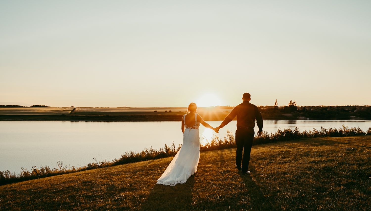A couple holds hands while walking along a lakeside path during a golden sunset.