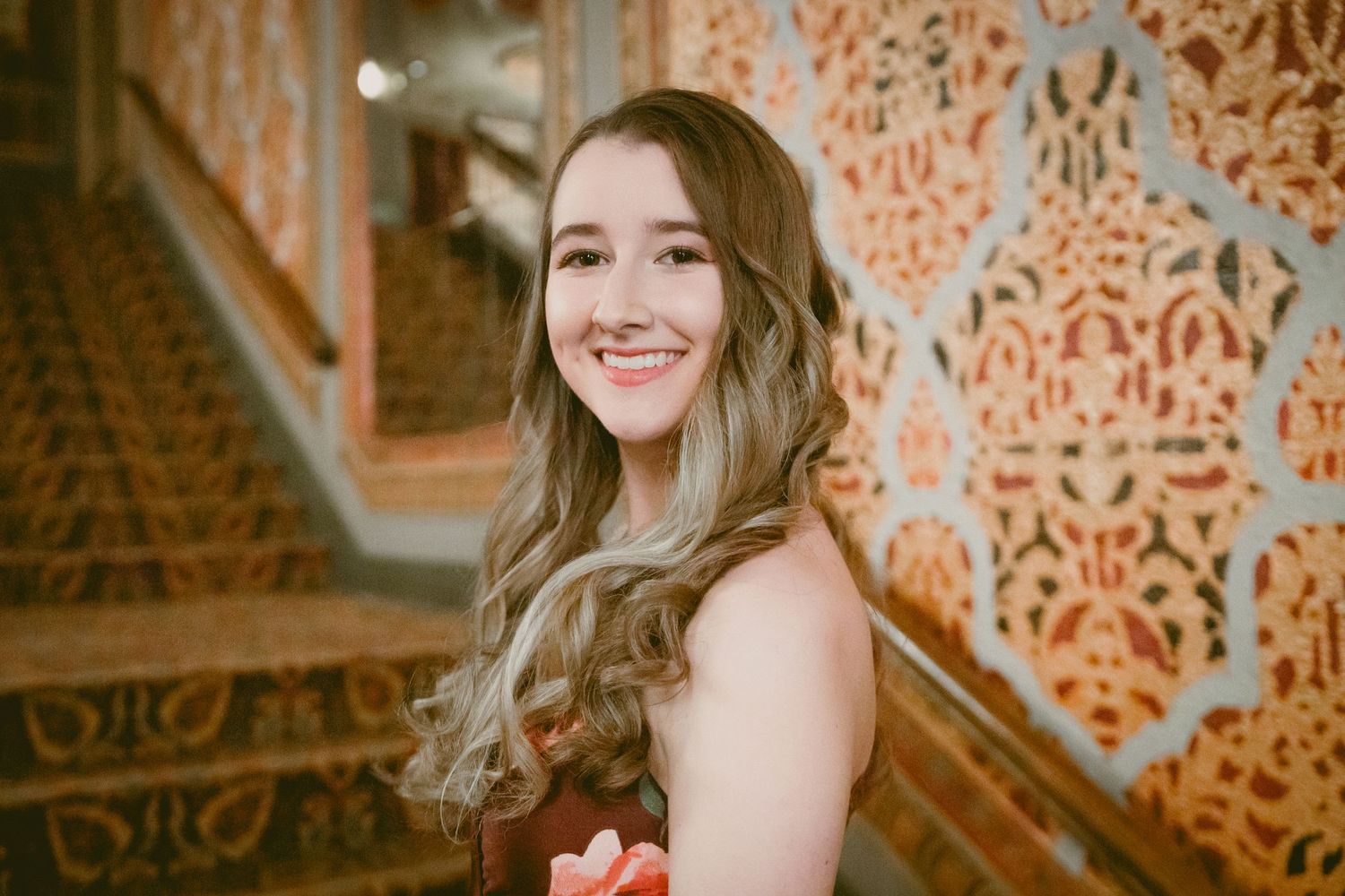Woman in formal wear smiling against decorative patterned wall background.