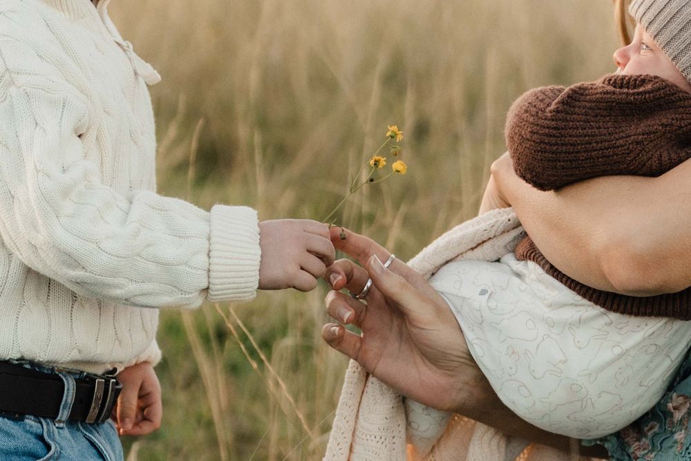 Family in a field, child holding wildflowers, parents embracing in cozy knit sweaters during a warm autumn day.