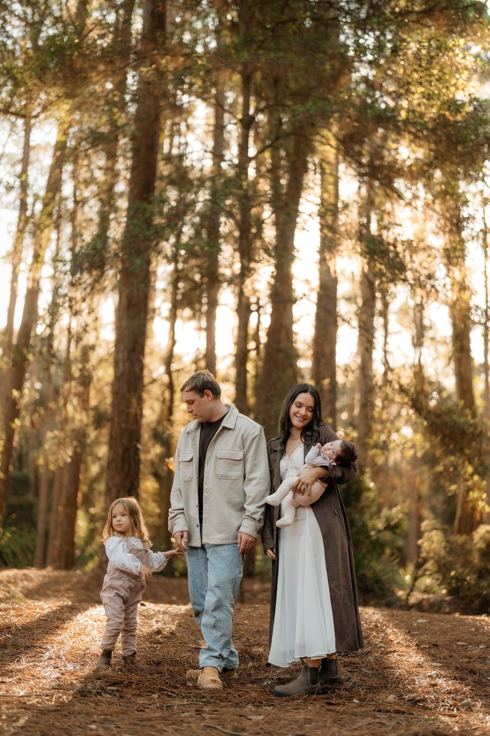 Family of four walking on a forest path at golden hour, parents holding toddler's hand while mom cradles a baby.