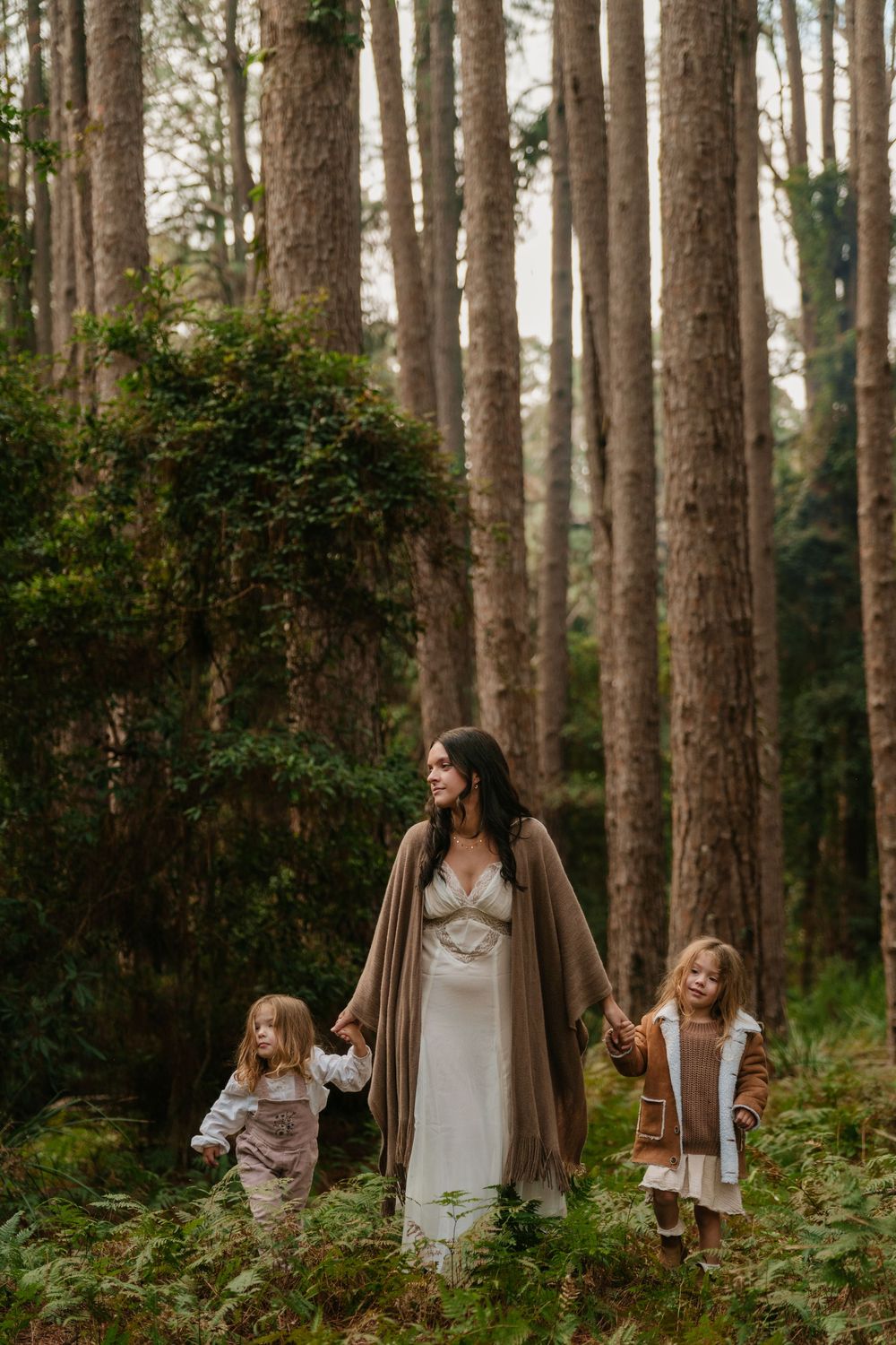 A woman in a white dress walks through a forest holding hands with two young children among tall trees and ferns.