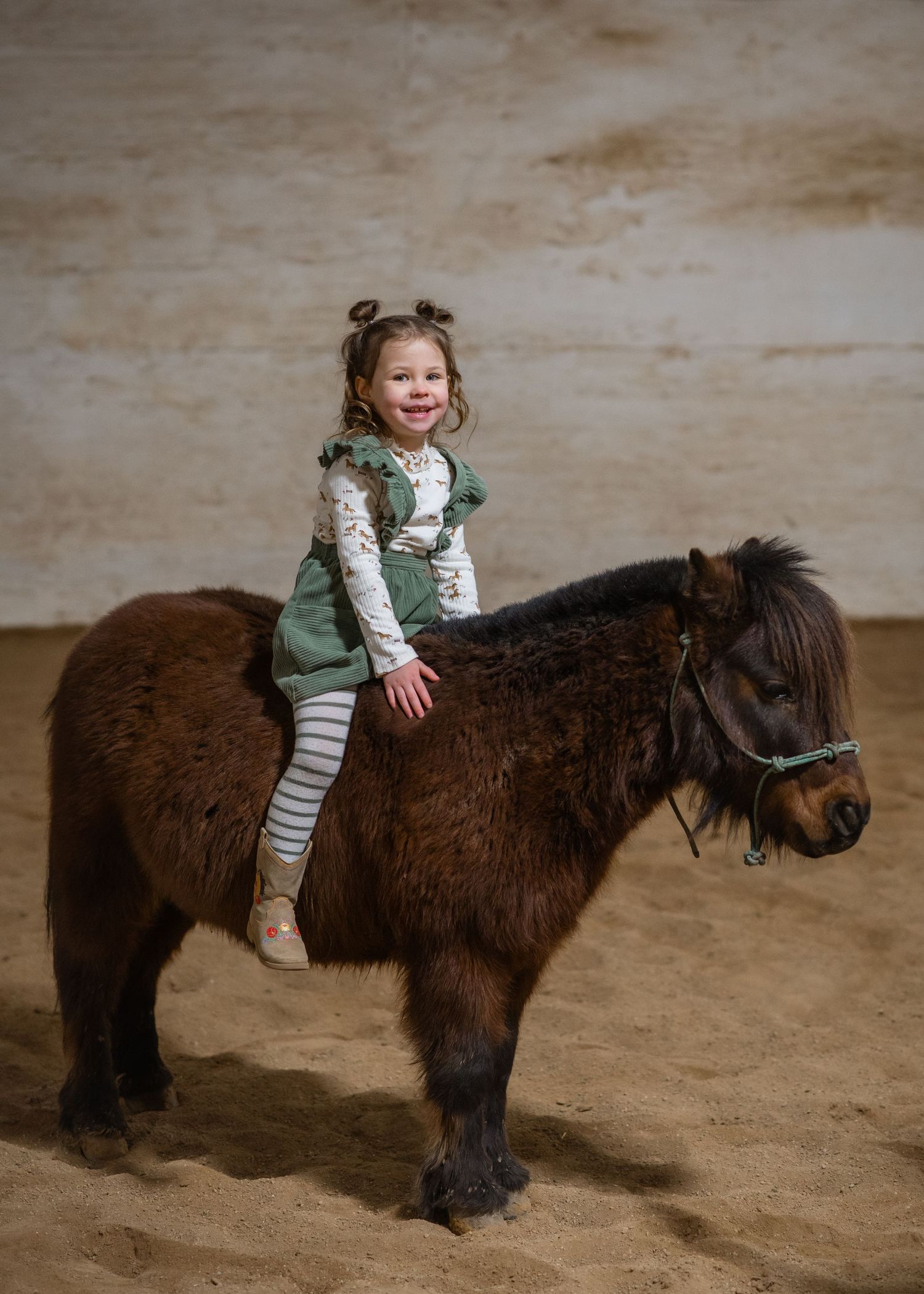 Small child riding a fluffy dark brown pony while wearing a green outfit with striped pants.