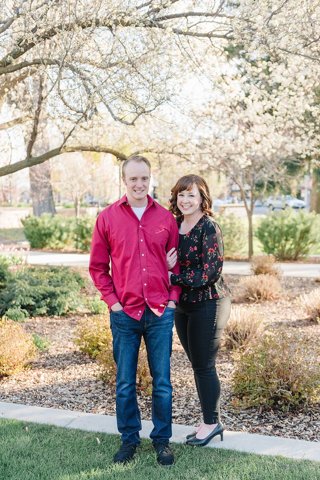 A couple stands together on a garden path surrounded by blooming spring trees.