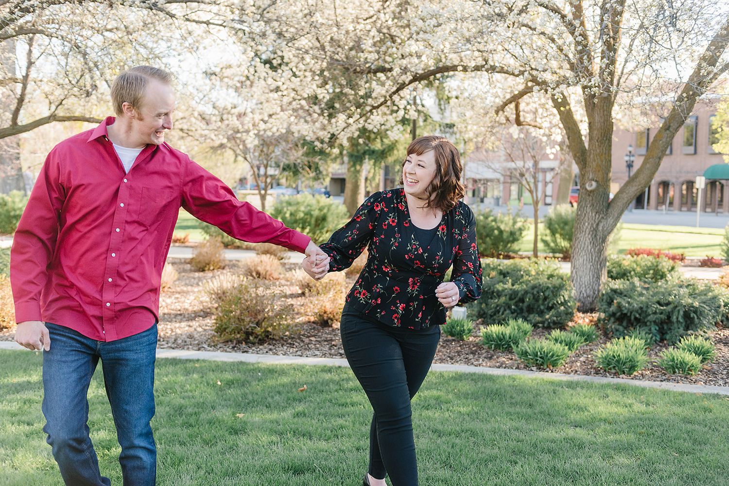 A couple dances playfully on a green lawn surrounded by blooming spring trees.