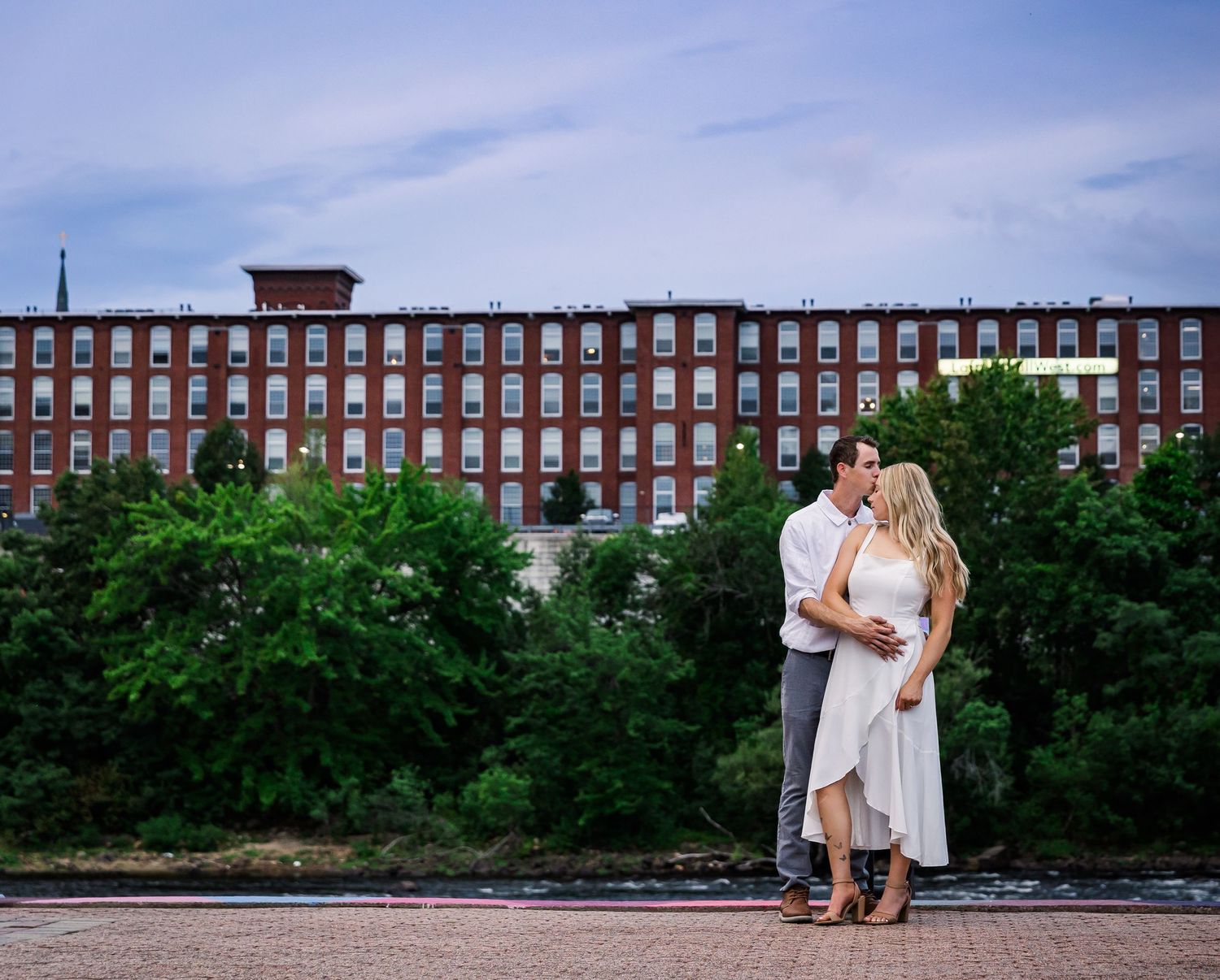 A romantic couple embraces in front of a historic brick factory building on a summer evening.