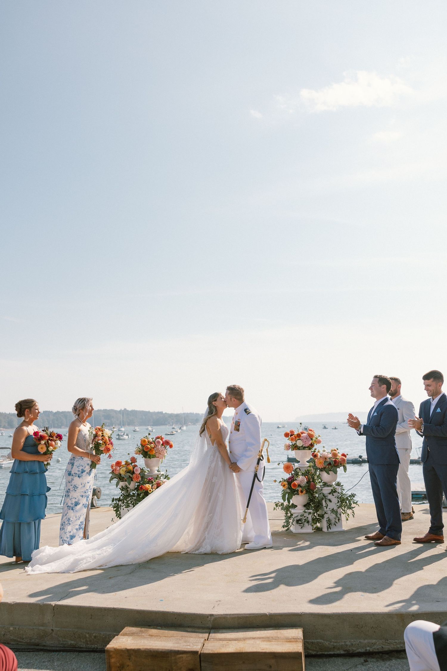 Wedding party lined up on stone ledge overlooking harbor with floral arrangements.