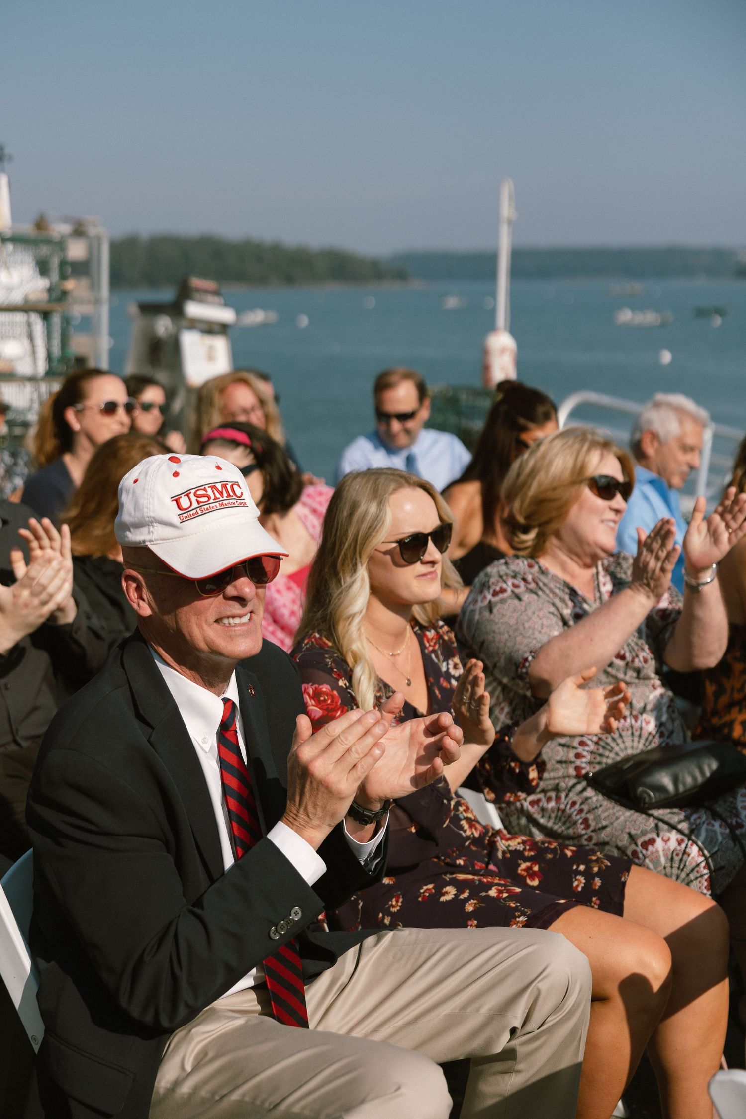 Wedding guests watching ceremony by the water on a sunny afternoon.