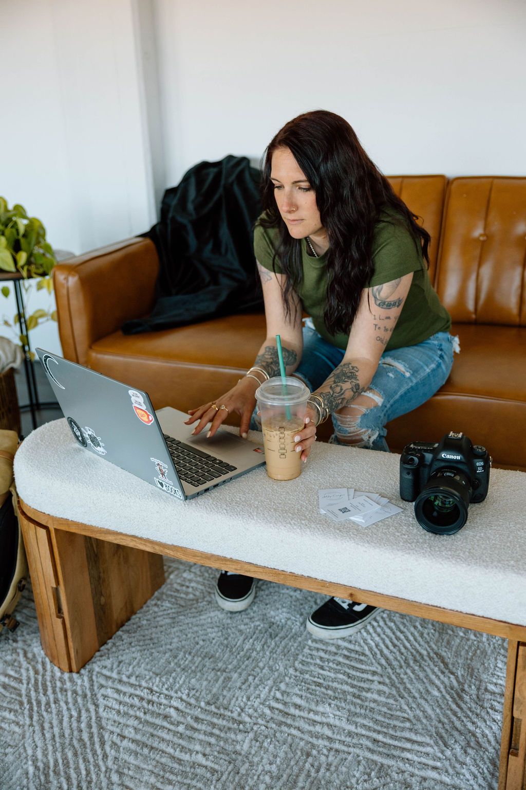 Person working on laptop while relaxing on brown leather couch with coffee and camera nearby.