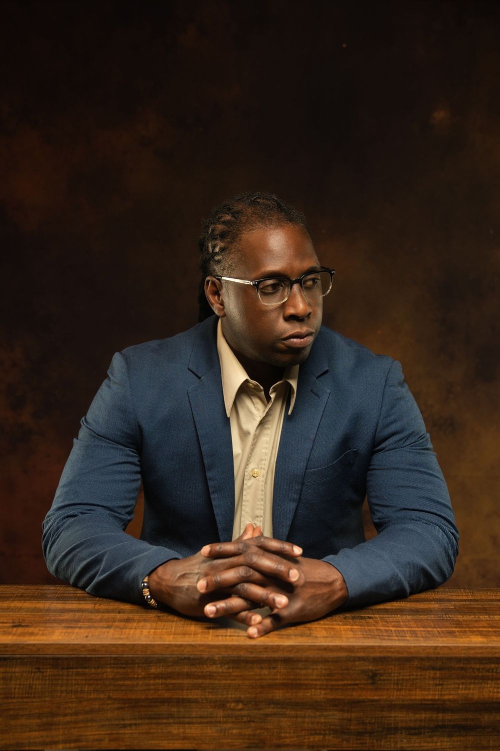 Professional Black man in blue blazer and glasses sits at wooden table with hands clasped against dark brown background.