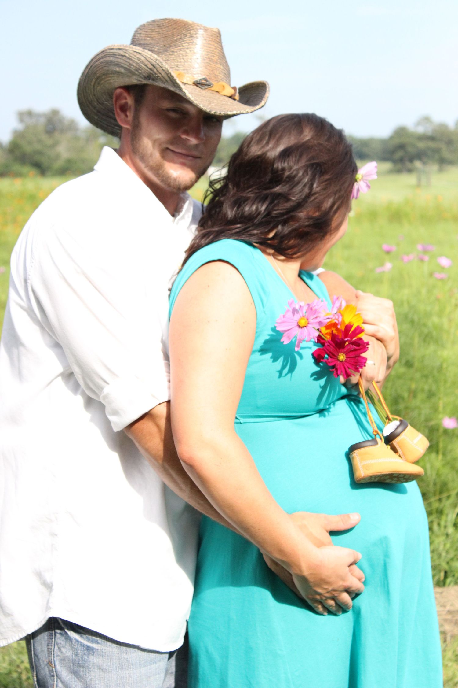 A couple in a turquoise dress and white shirt embrace outdoors in a sunny field with pink flowers.
