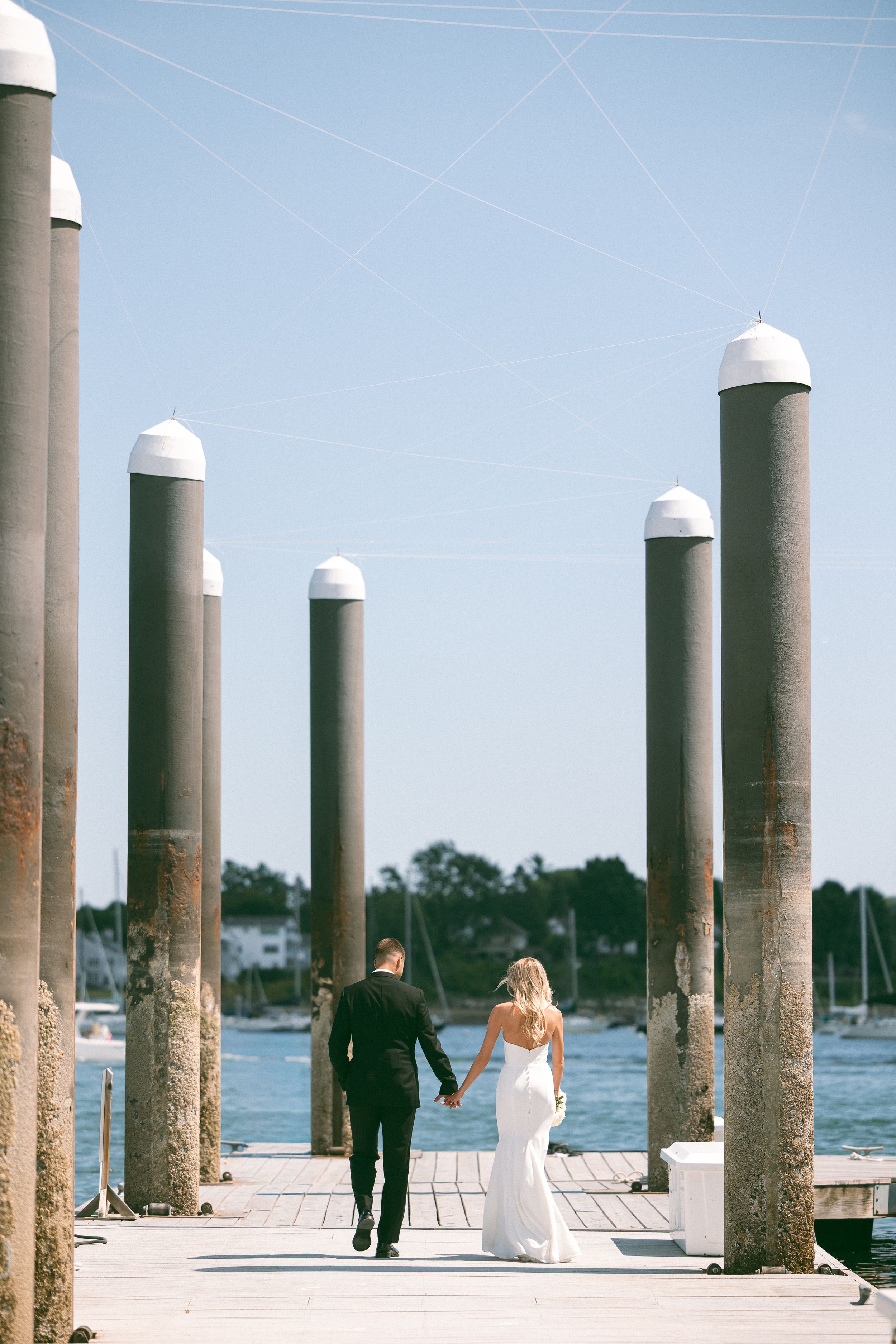 A bride and groom walk hand in hand along a sunlit marina dock, surrounded by tall pillars with a blue sky backdrop.