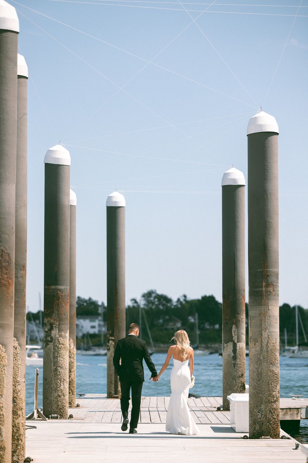 A bride and groom walk hand in hand along a sunlit marina dock, surrounded by tall pillars with a blue sky backdrop.