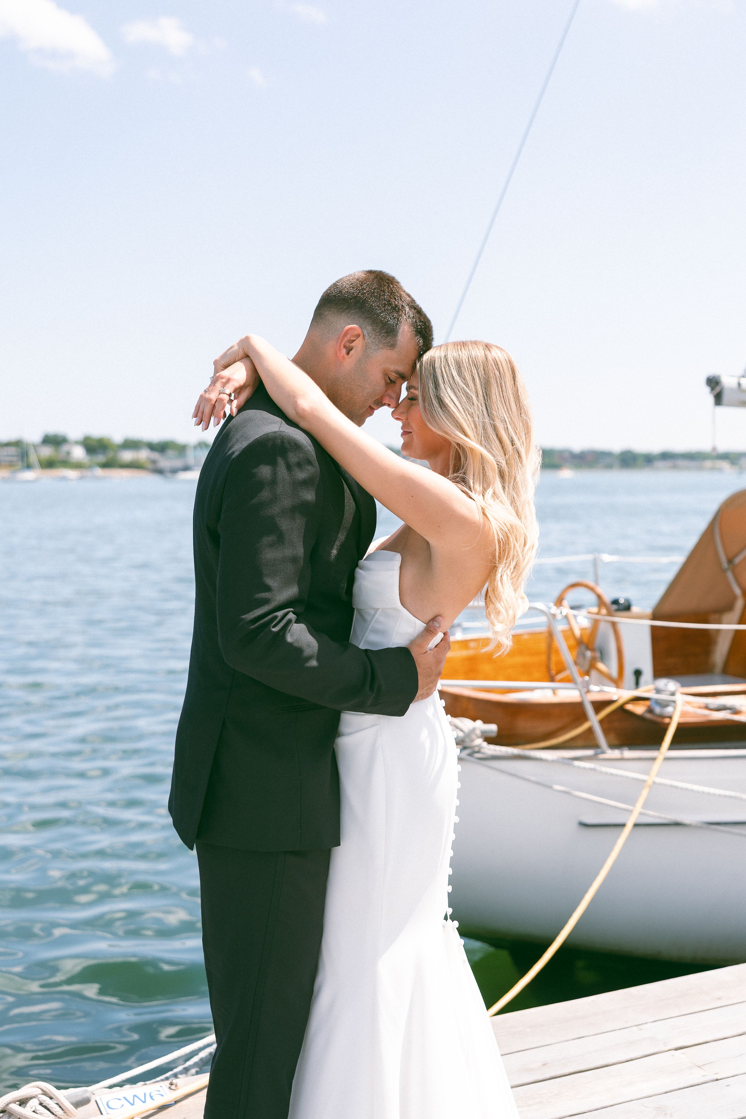 Bride and groom embrace on a sunny dock beside a sailboat, sharing a romantic moment in wedding attire.