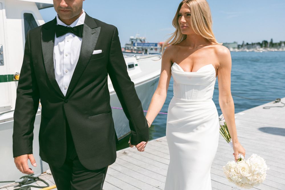 Bride and groom holding hands on a waterfront dock, bride in strapless white gown holding bouquet, groom in black tuxedo.