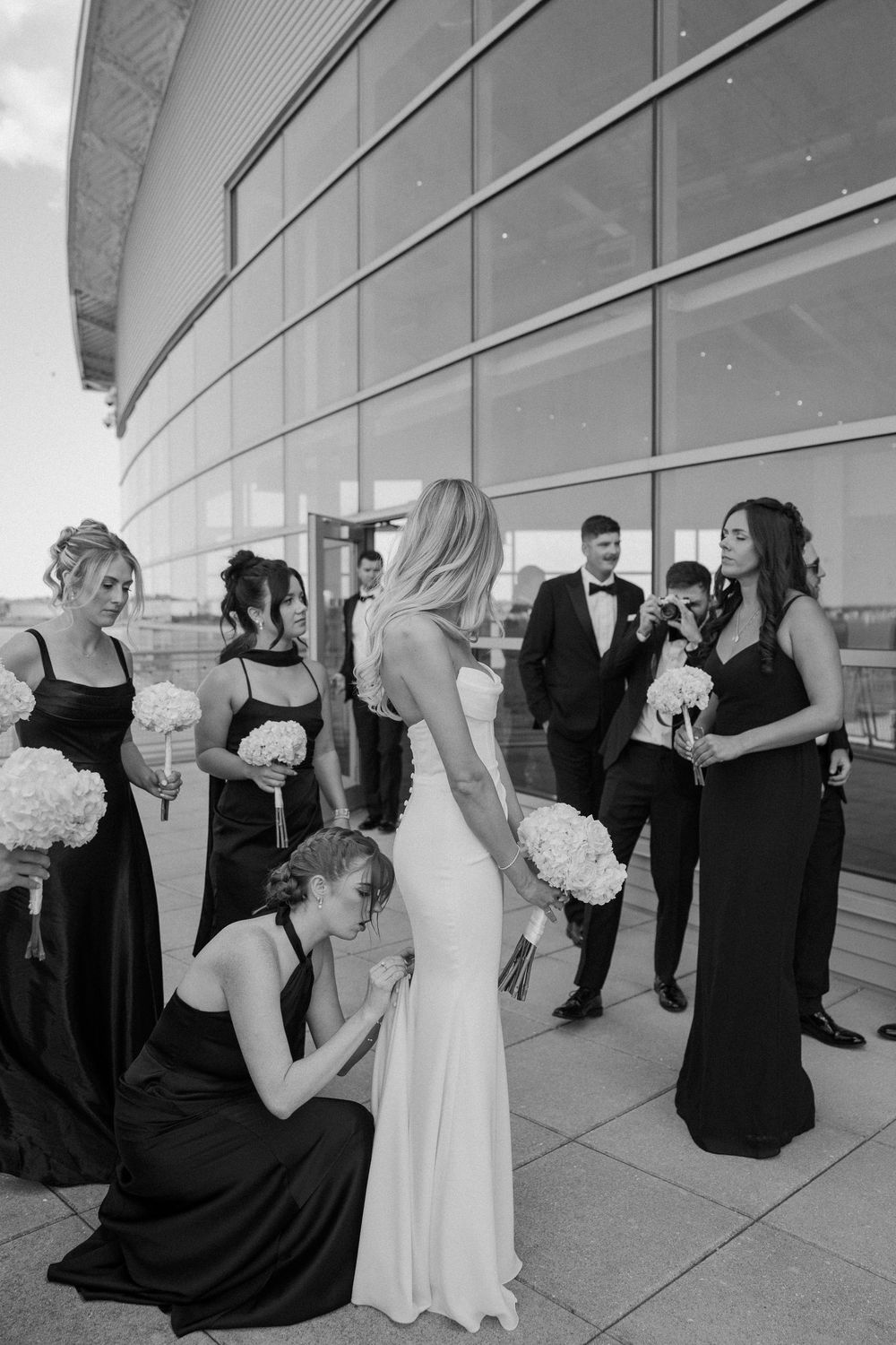 Black and white wedding photo of bride in white gown with bridesmaids in black dresses holding white bouquets outside modern building.