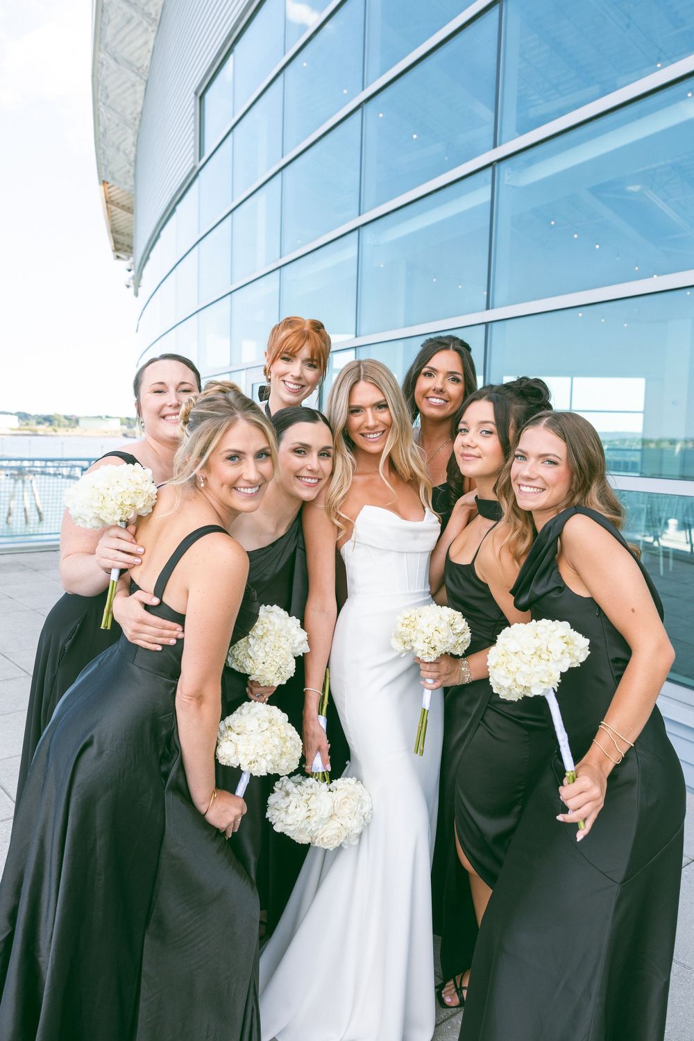 Bride in white gown poses with bridesmaids in black dresses holding white floral bouquets outside modern glass building.